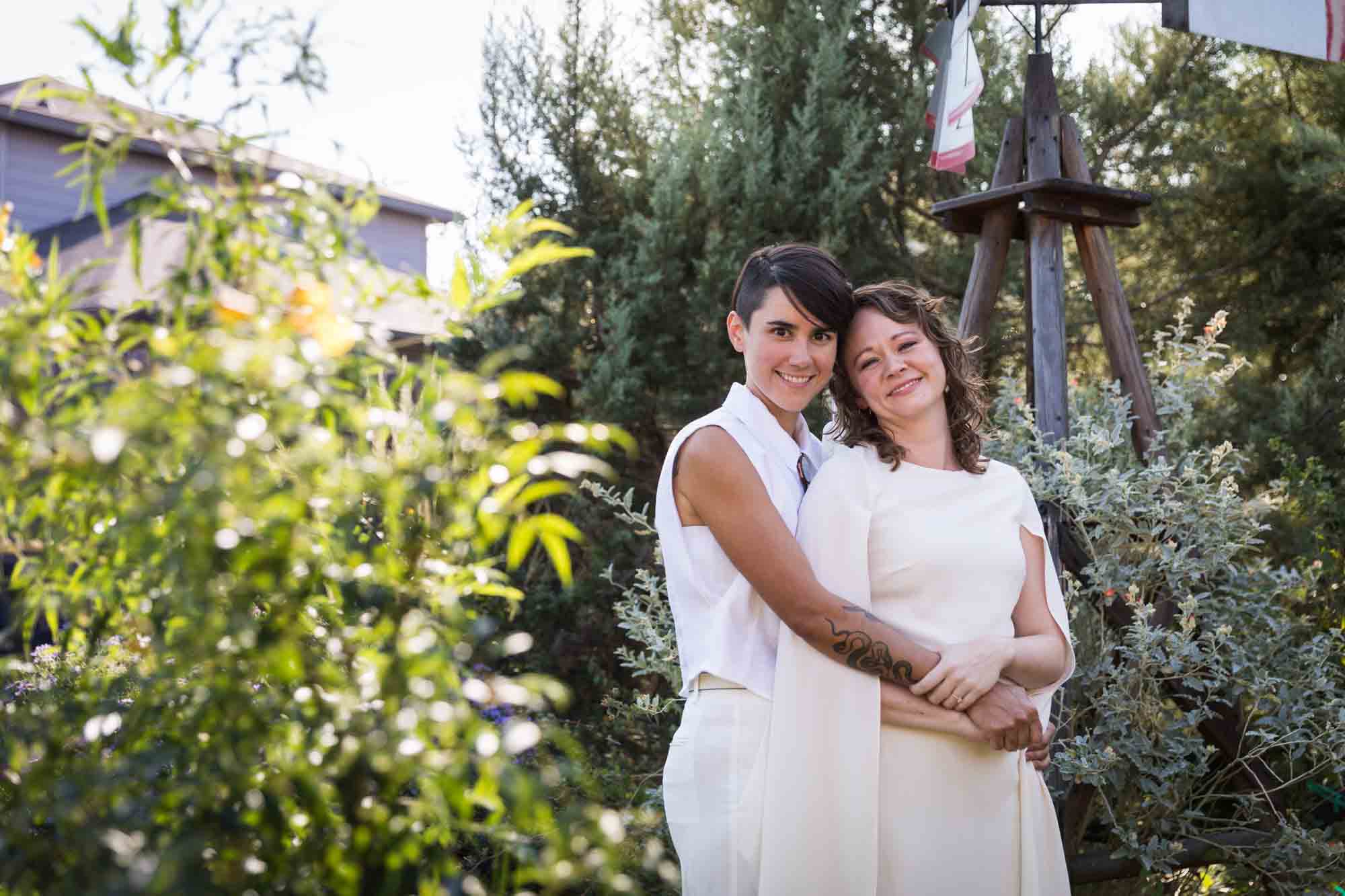 Two brides wearing white and hugging surrounded by plants at a Boerne backyard wedding