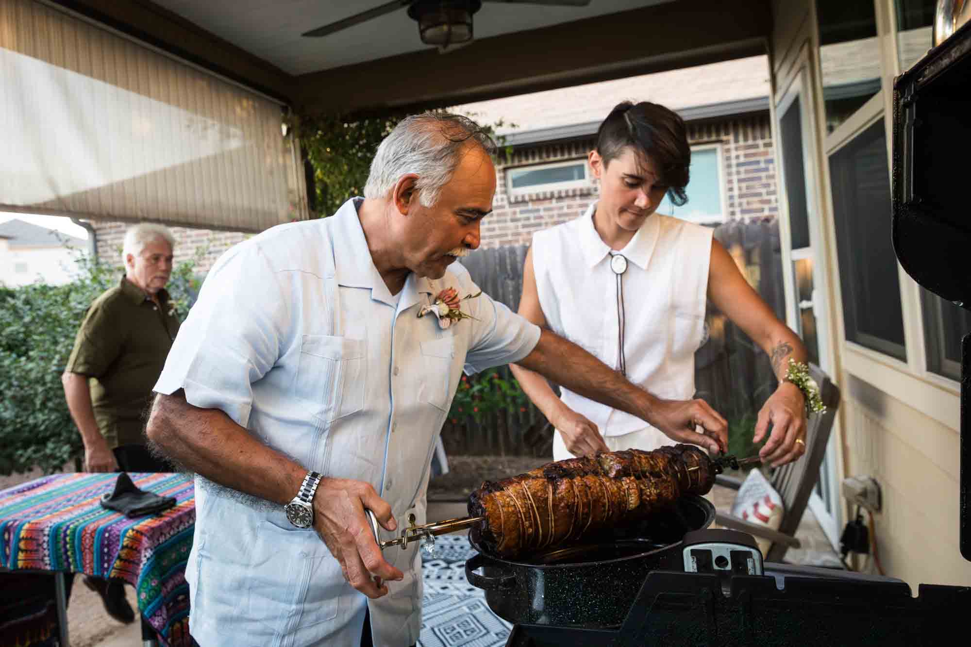 Father holding up pork loin on a spit over a grill with woman wearing sleeveless white shirt in the background