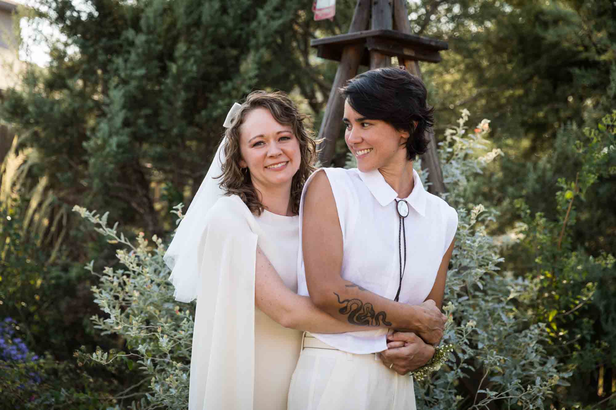 Two brides wearing white and hugging surrounded by plants at a Boerne backyard wedding