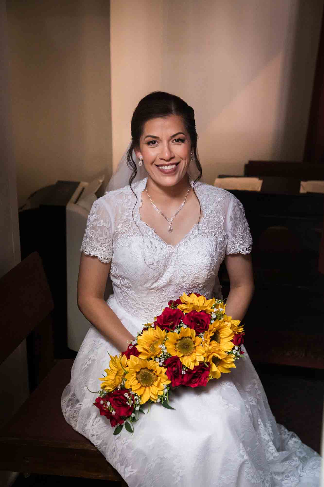 Bride sitting while holding bouquet of sunflowers and red roses during St. Henry Catholic Church wedding