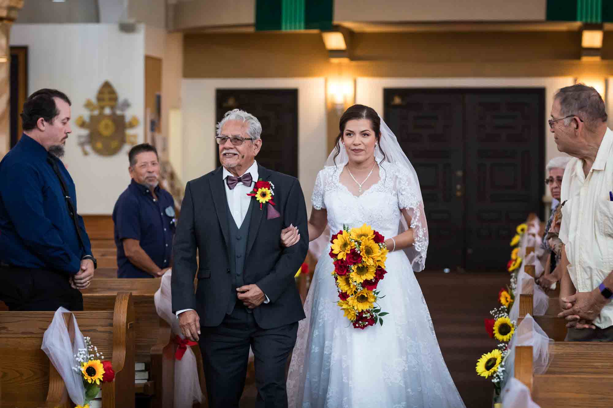 Bride holding father's arm while walking down aisle in front of guests during St. Henry Catholic Church wedding