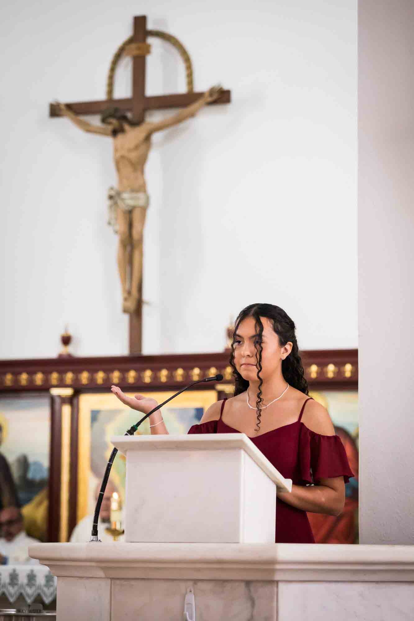 St. Henry Catholic Church wedding photos of bridesmaid wearing red dress and holding hand up in the air at podium with crucifix in background