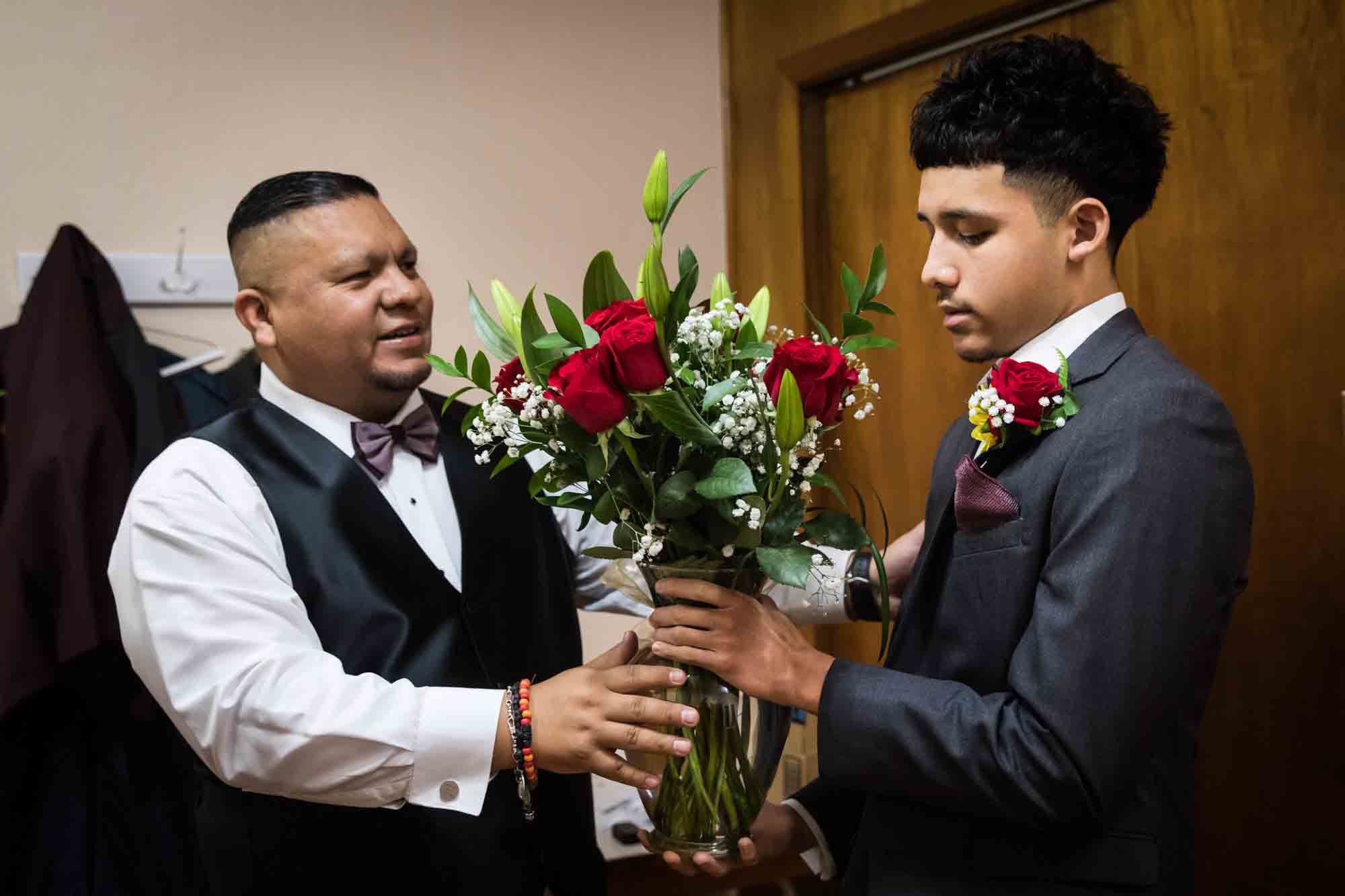 Father accepting vase of red roses from young man in room during St. Henry Catholic Church wedding