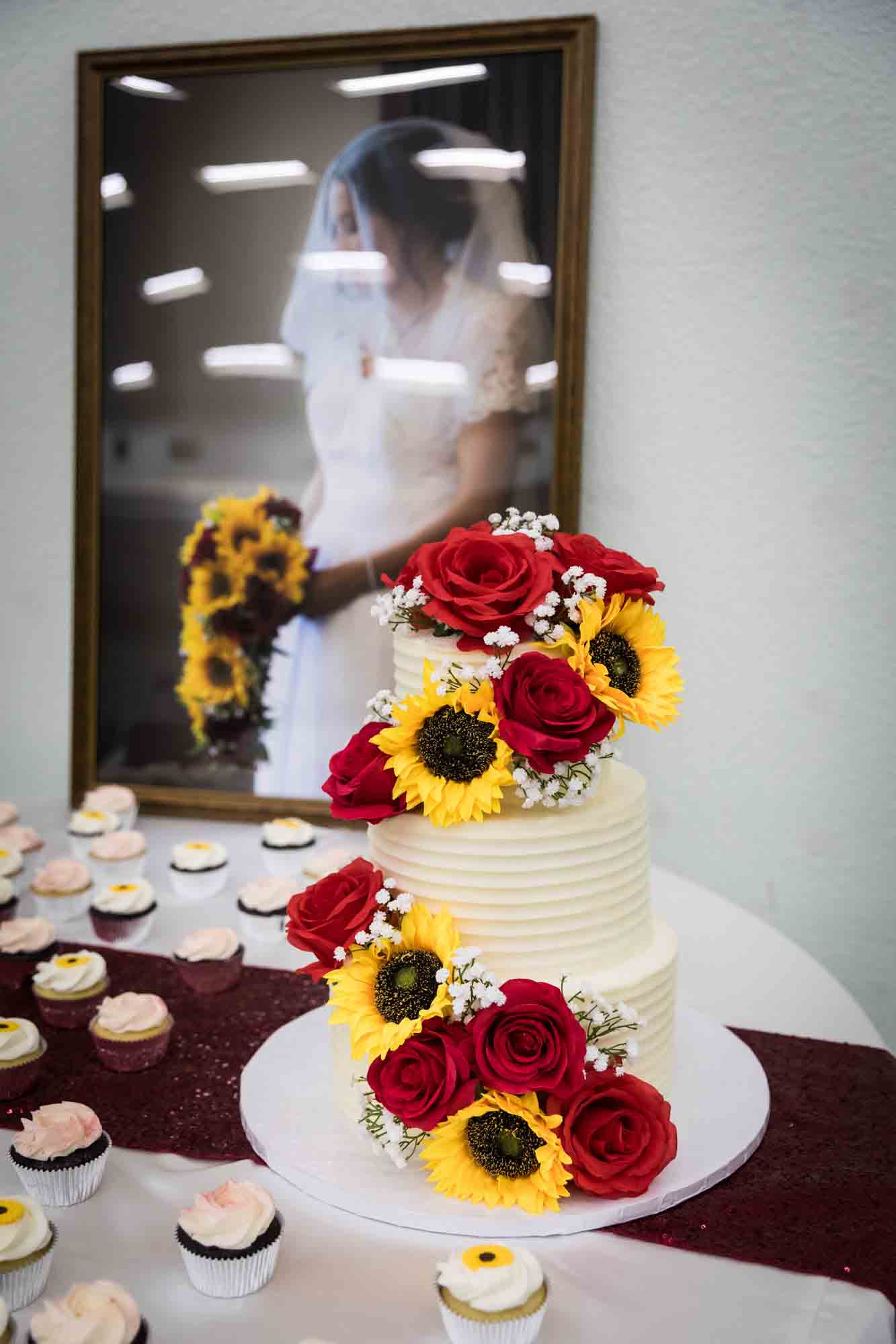 Wedding cake decorated with sunflowers and red roses in front of large framed photo of bride