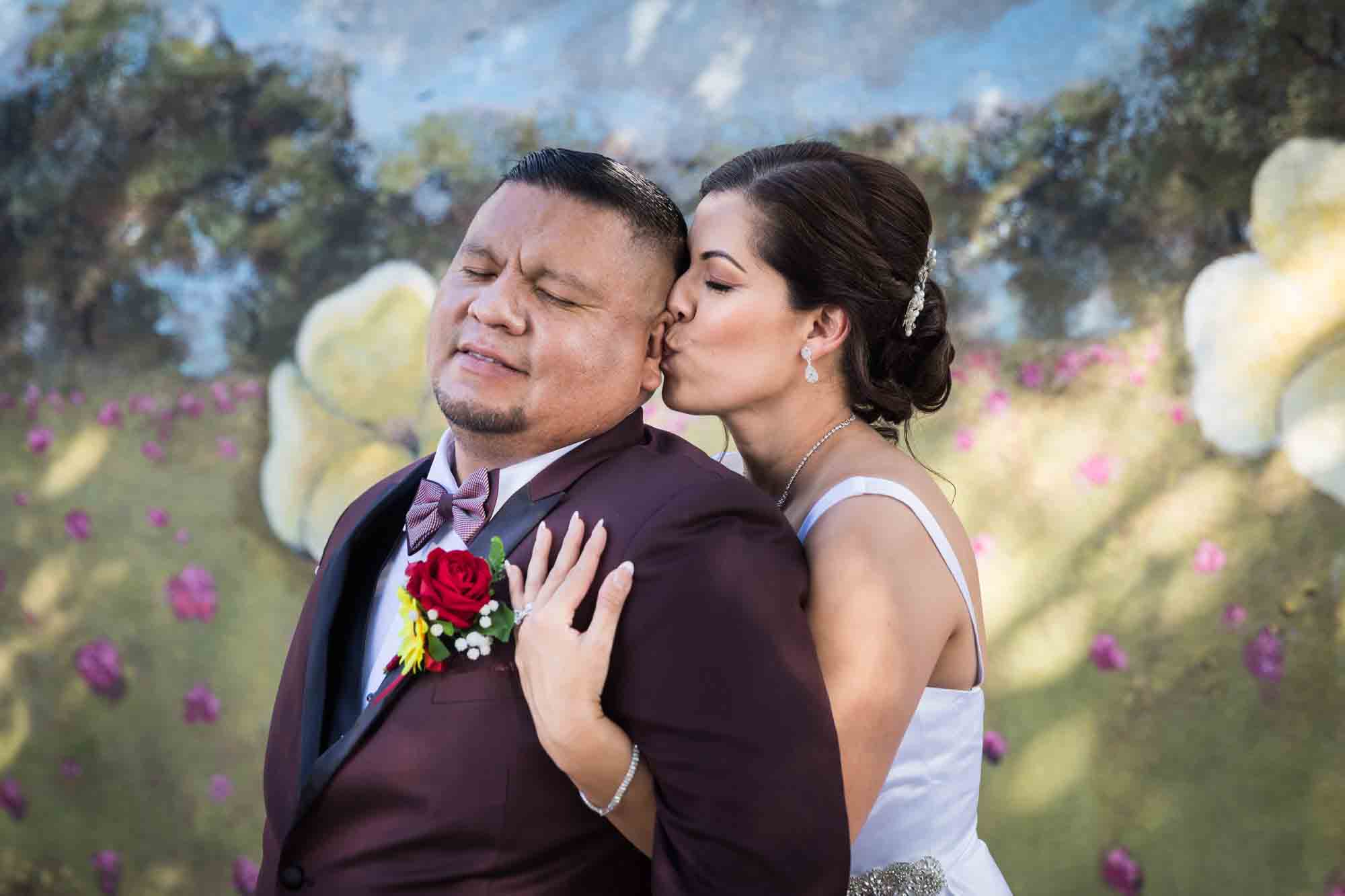 St. Henry Catholic Church wedding photos of bride kissing groom's ear in front of floral mural