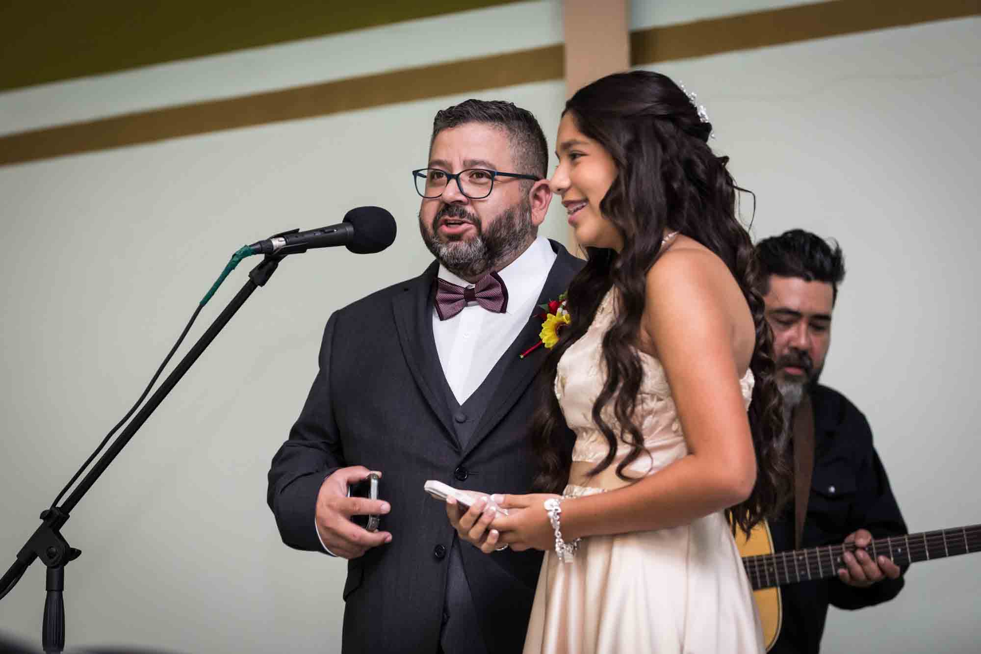 Man wearing glasses and bow tie with girl wearing off white dress talking into a microphone during a St. Henry Catholic Church wedding reception