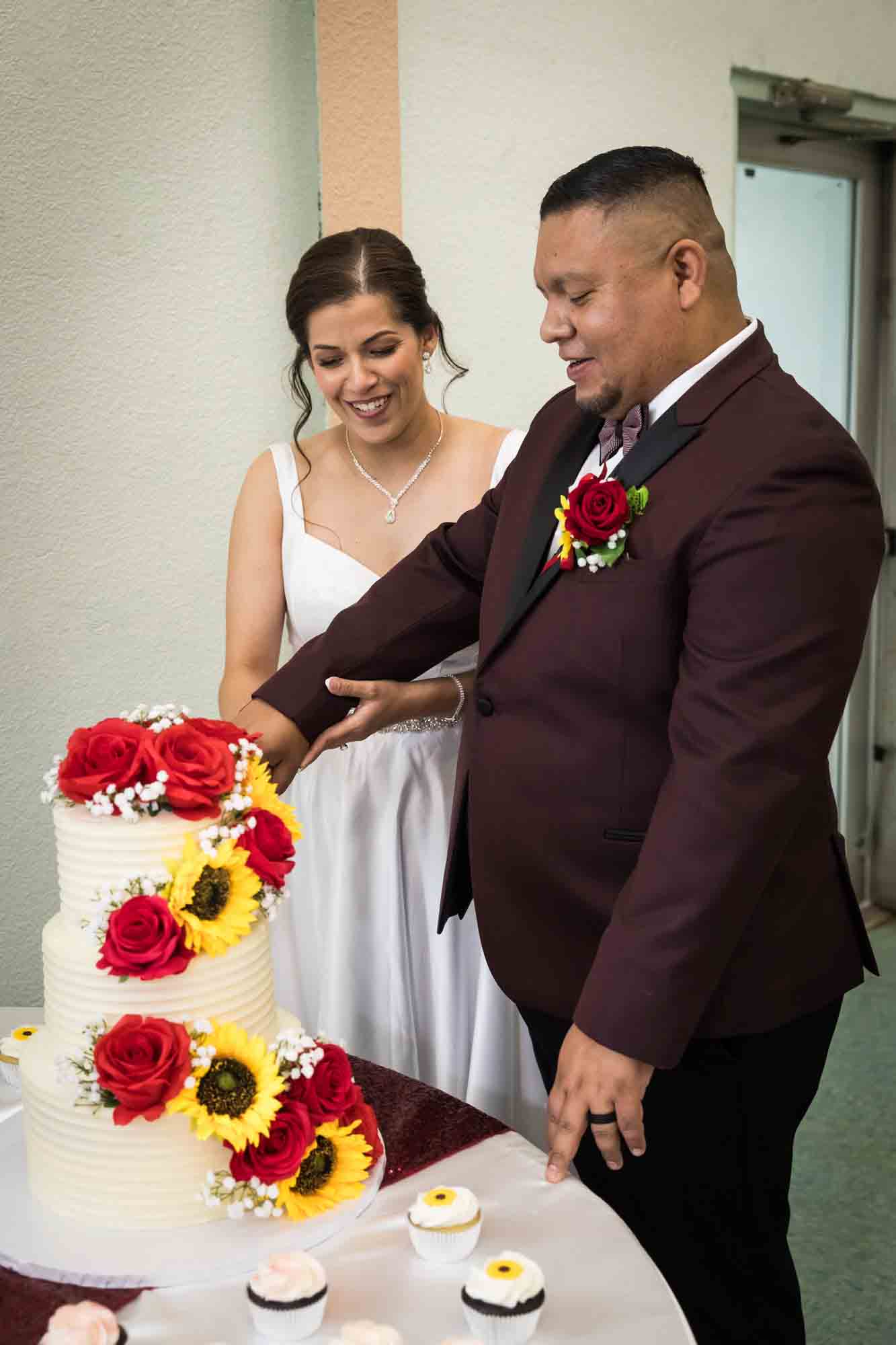 Bride and groom cutting wedding cake decorated with sunflowers and red roses during a St. Henry Catholic Church wedding reception