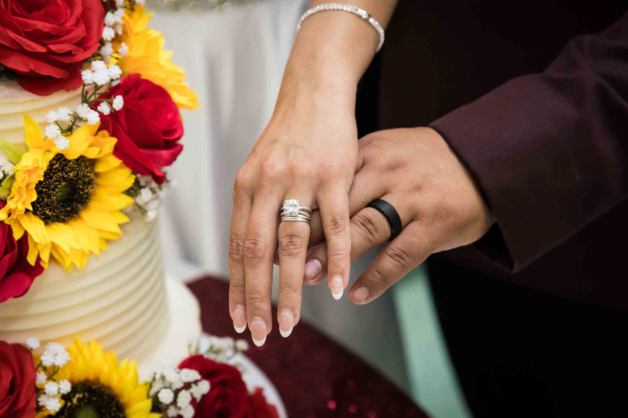 Close up of bride and groom showing wedding rings in front of wedding cake decorated with sunflowers and red roses