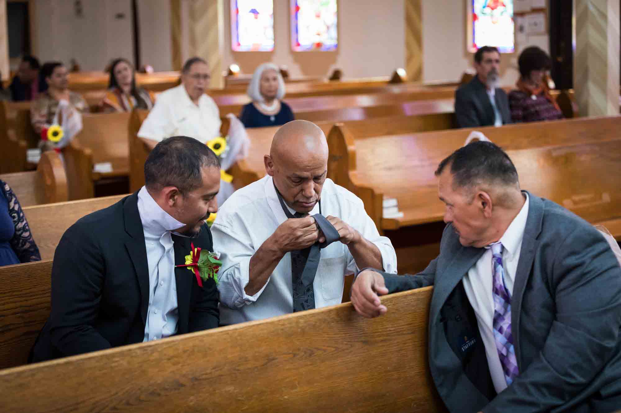 Two men watching another man put on a tie while sitting in pew during St. Henry Catholic Church wedding