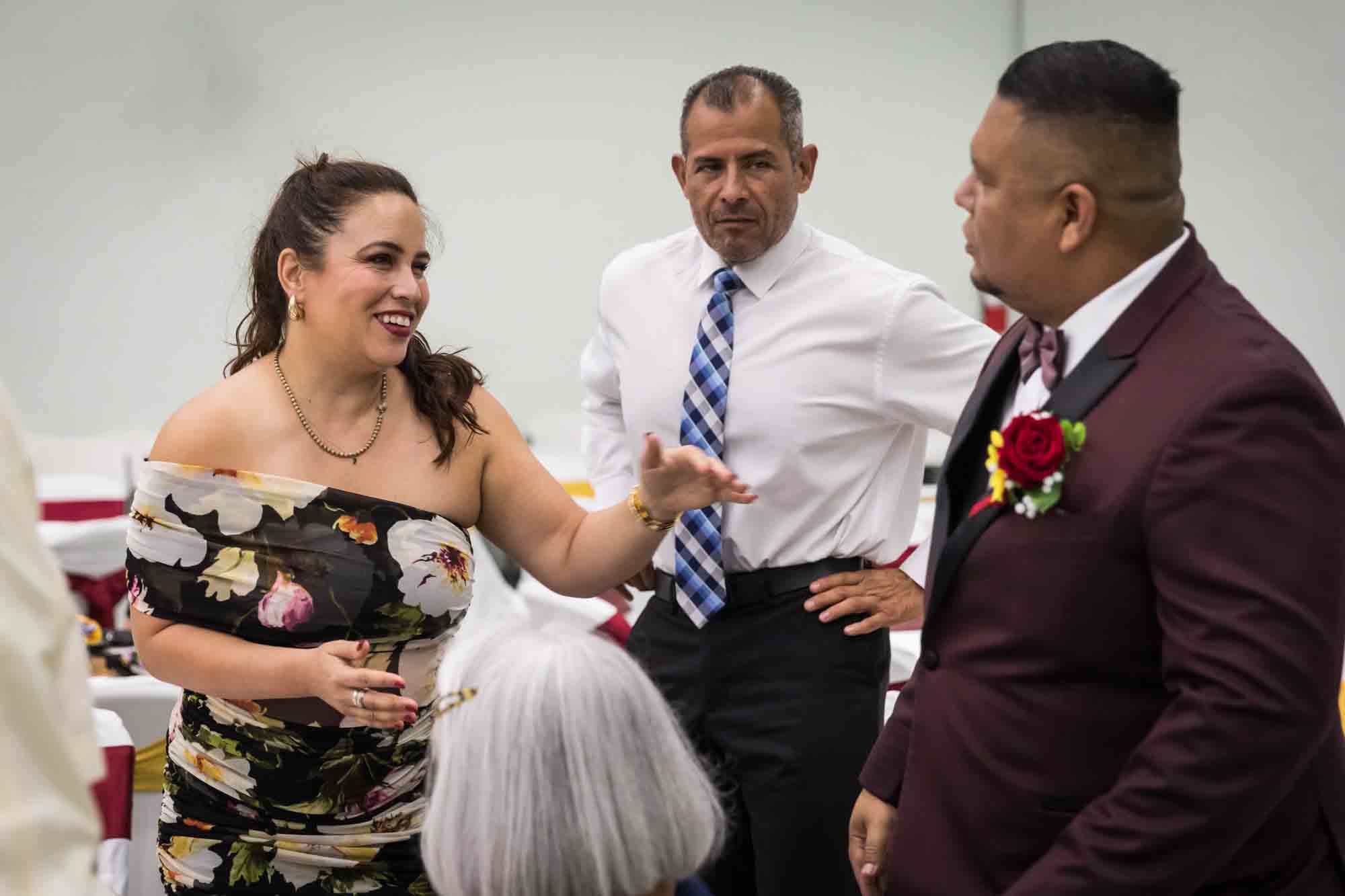 Groom talking to woman wearing off shoulder, floral dress and man wearing tie during a St. Henry Catholic Church wedding reception