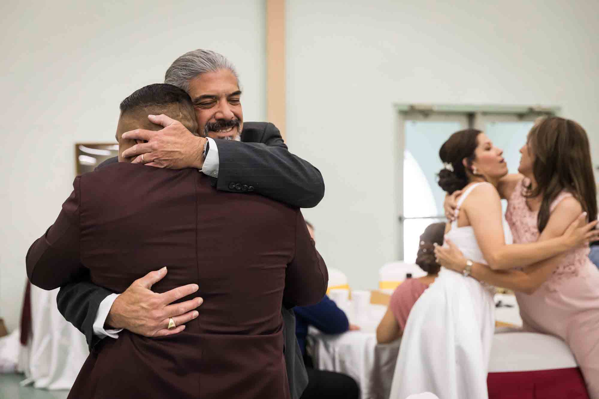 Groom hugging man with gray hair with bride hugging woman in background during a St. Henry Catholic Church wedding reception