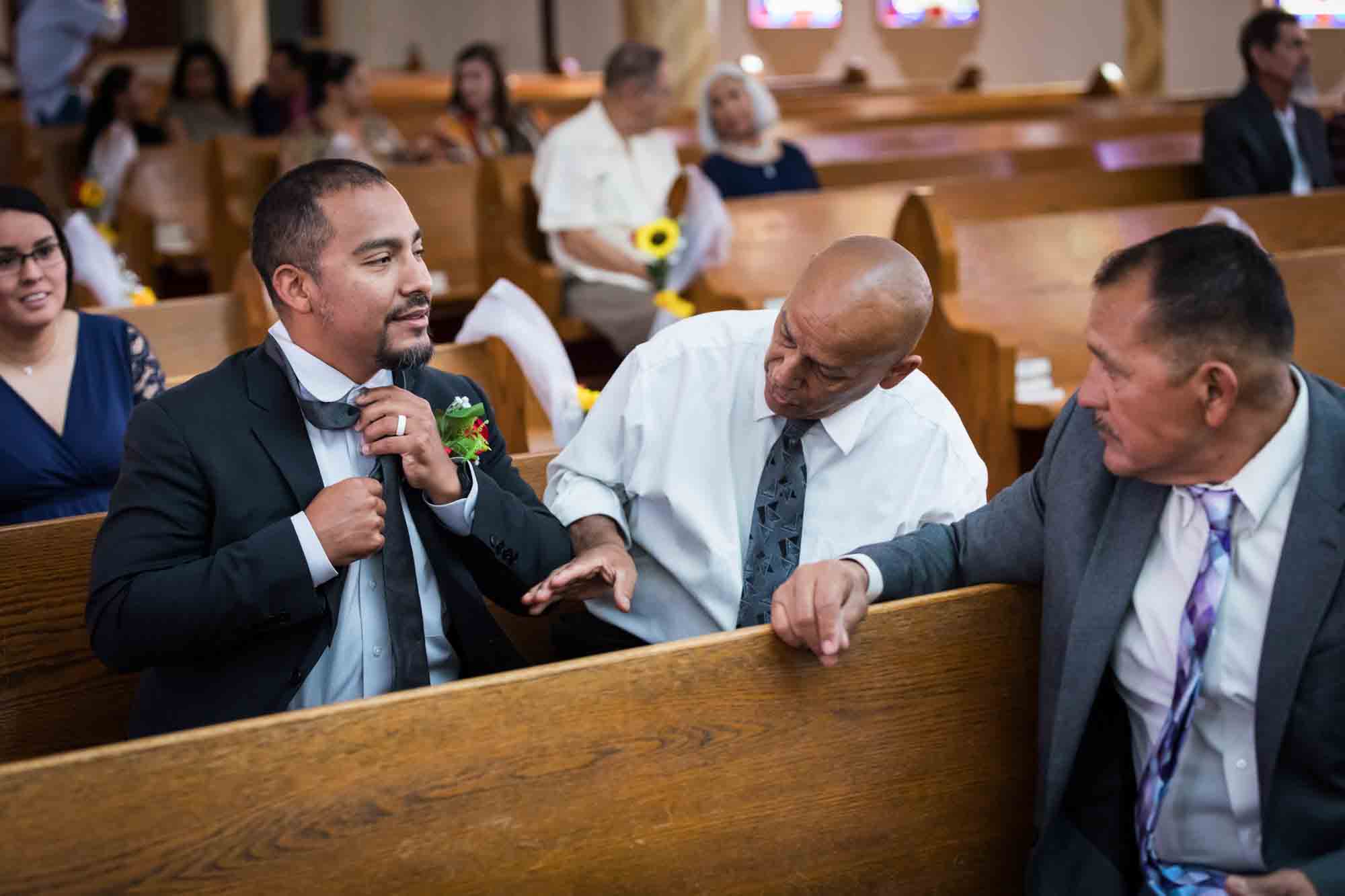 Two men sitting in pew showing man how to put on tie during St. Henry Catholic Church wedding