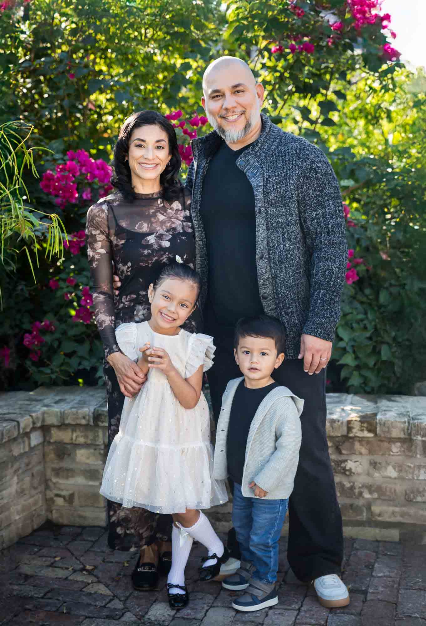 Parents standing with little boy and girl in front of brick wall and bushes during a La Villita family portrait session