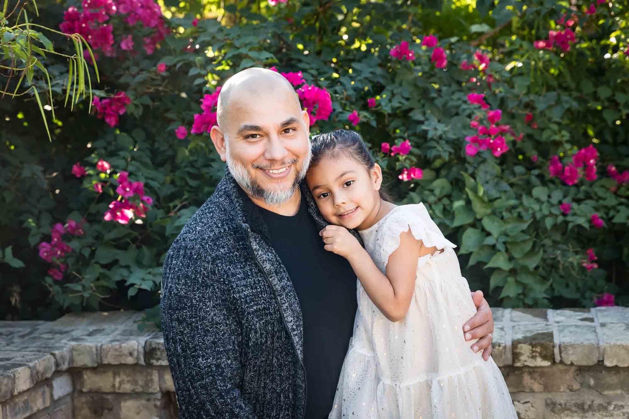 Father hugging little girl wearing white dress in front of bushes during a La Villita family portrait session