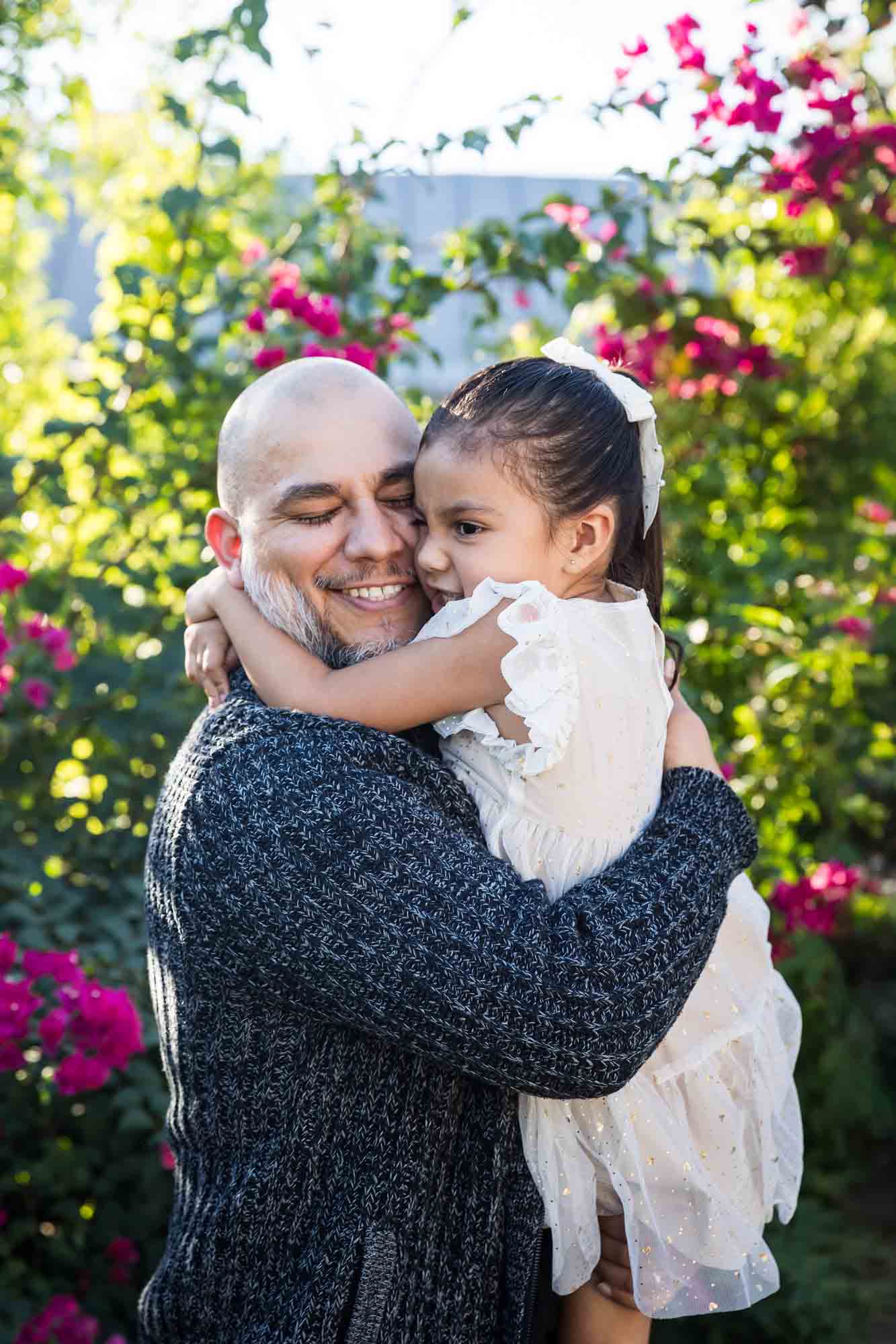 Father hugging little girl wearing white dress in front of bushes during a La Villita family portrait session