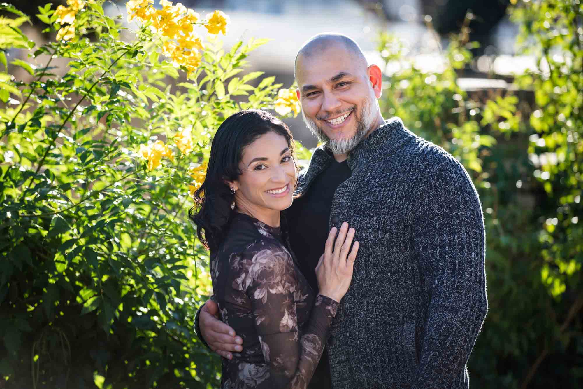 Couple hugging in front of bushes with yellow flowers during a La Villita family portrait session