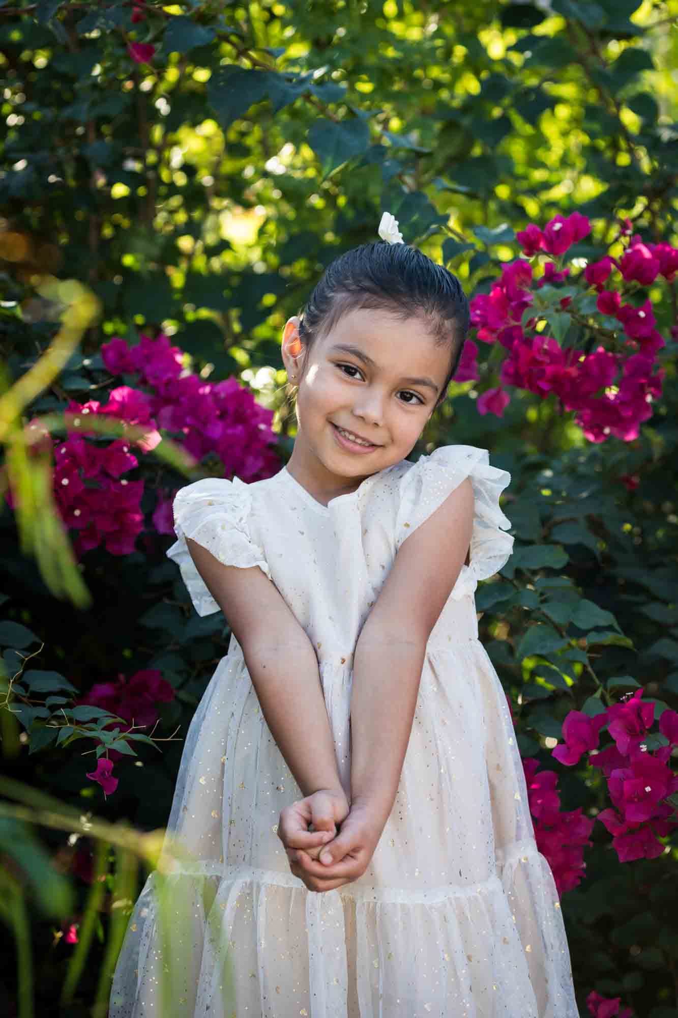 Little girl wearing white dress holding hands in front of self in front of brick wall and bushes during a La Villita family portrait session