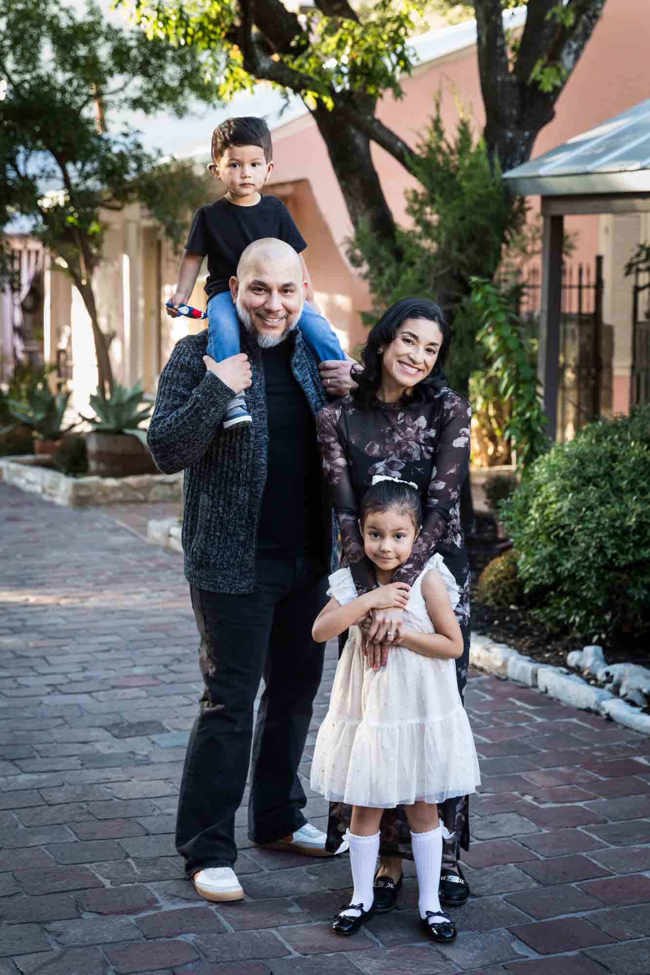 Father with little boy on his shoulder standing with mother and little girl on cobblestone street during a La Villita family portrait session