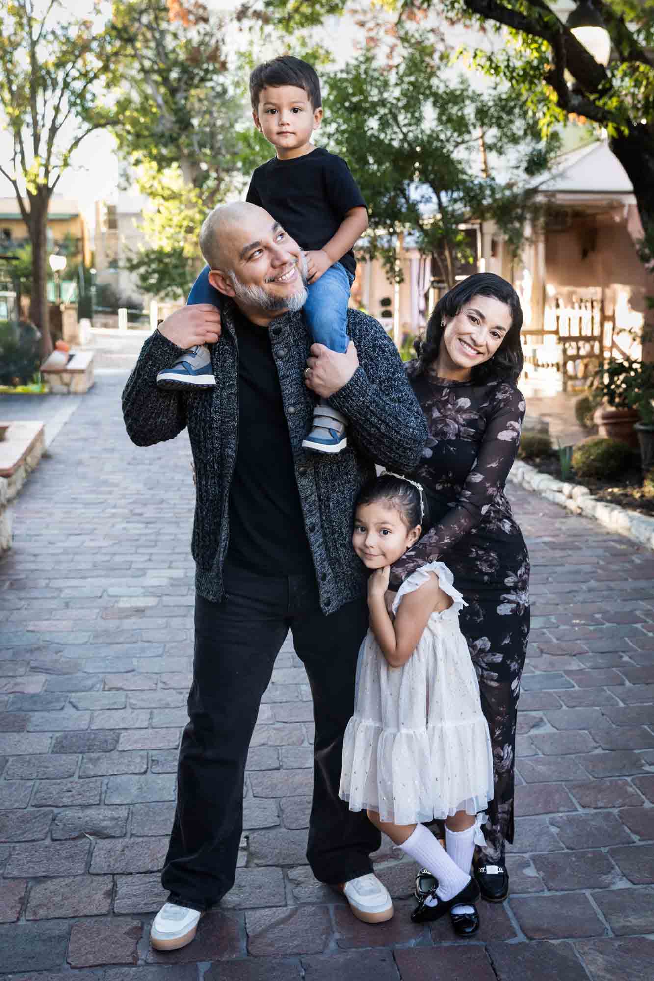 Father with little boy on his shoulder standing with mother and little girl on cobblestone street during a La Villita family portrait session