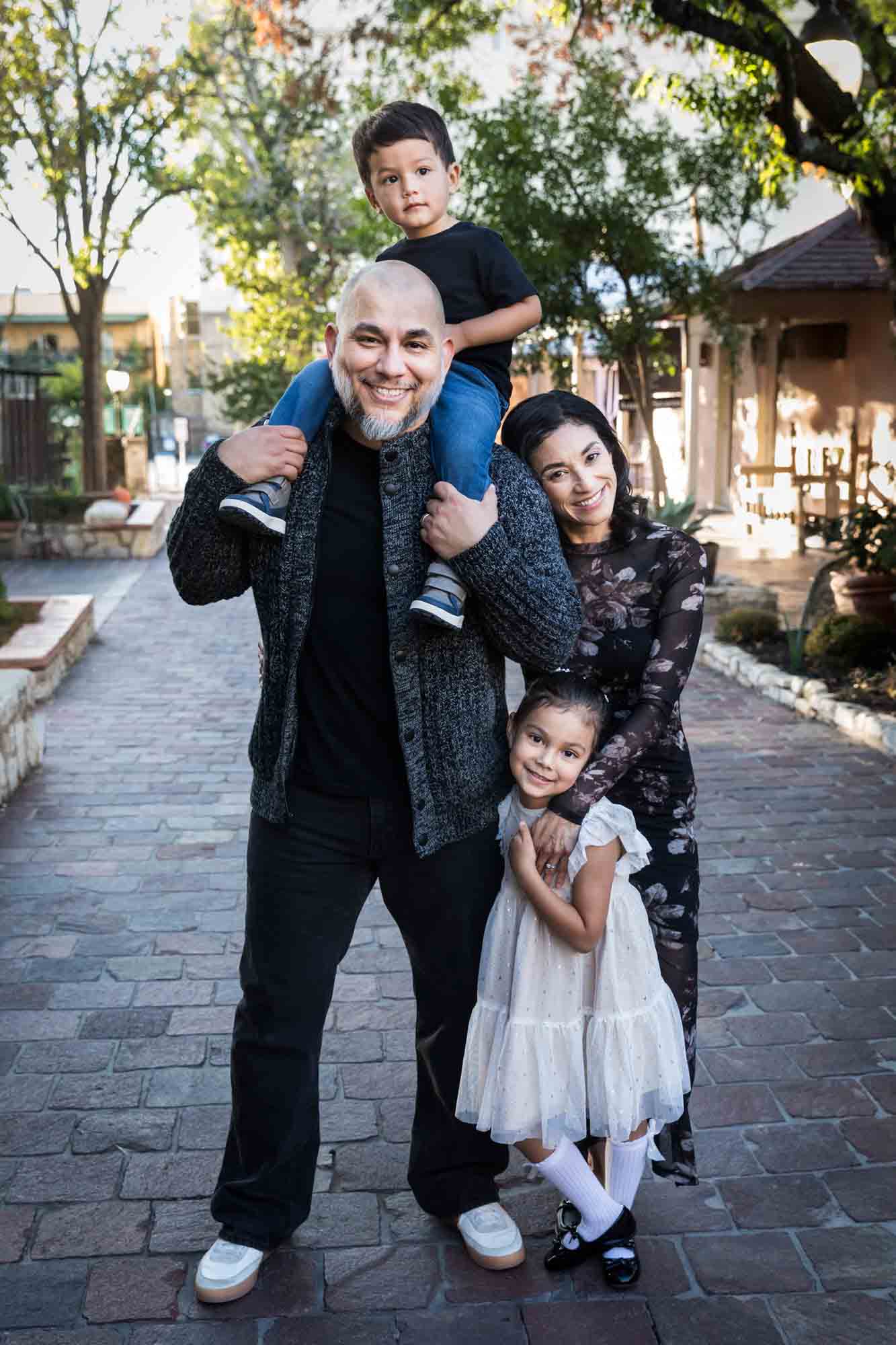 Father with little boy on his shoulder standing with mother and little girl on cobblestone street during a La Villita family portrait session