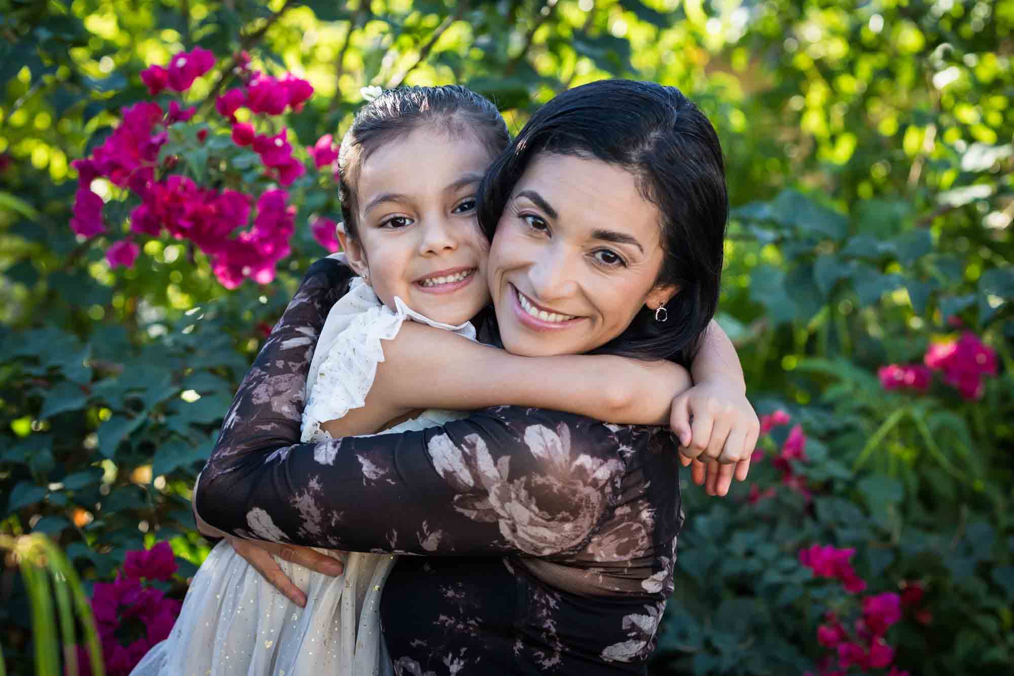 Mother hugging little girl wearing white dress in front of bushes during a La Villita family portrait session