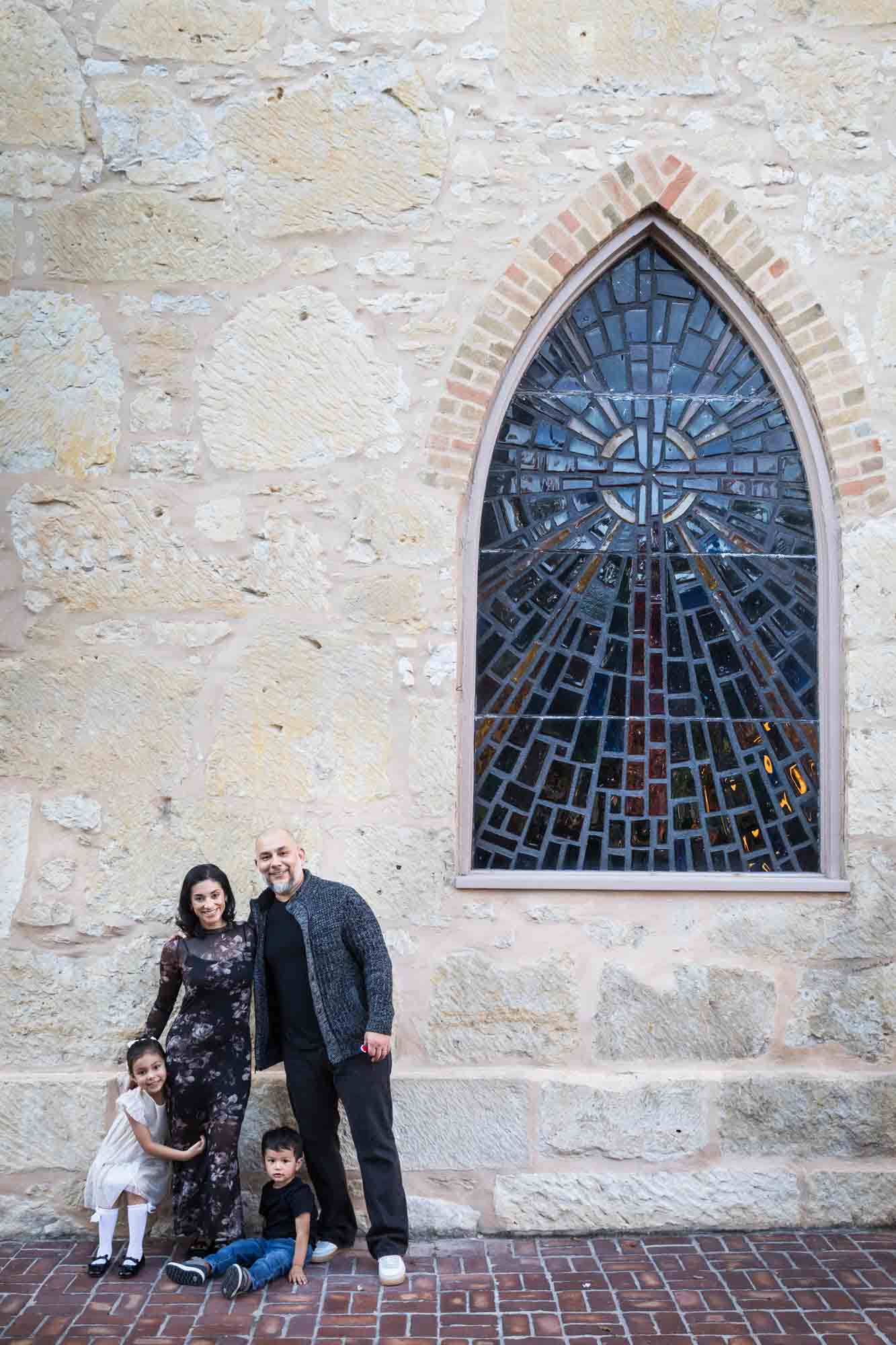 Parents and two young children standing to left of stained glass window against stone wall of Little Church during a La Villita family portrait session