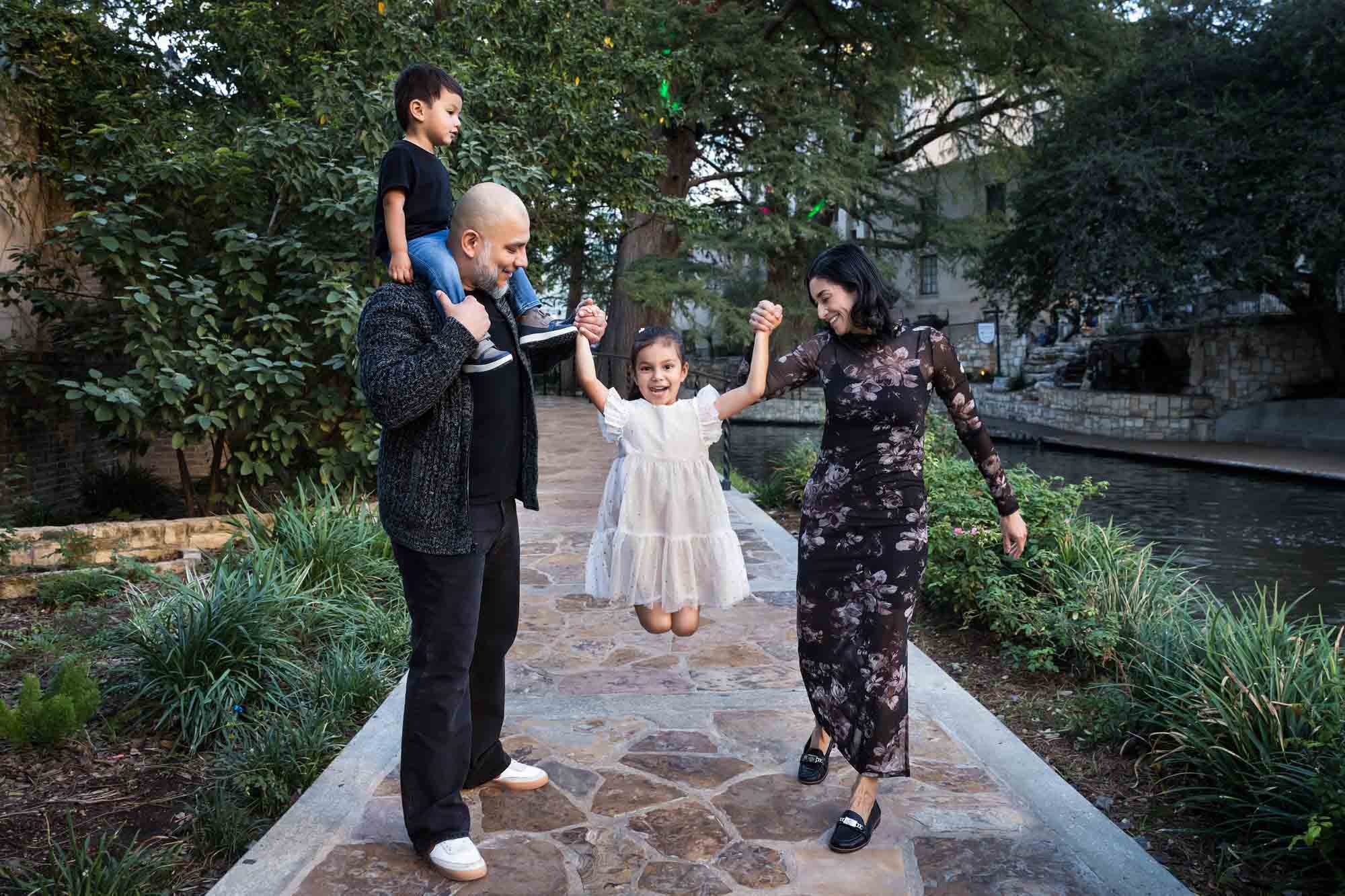 Father with little boy on his shoulders and mother holding up little girl wearing dress on the Riverwalk