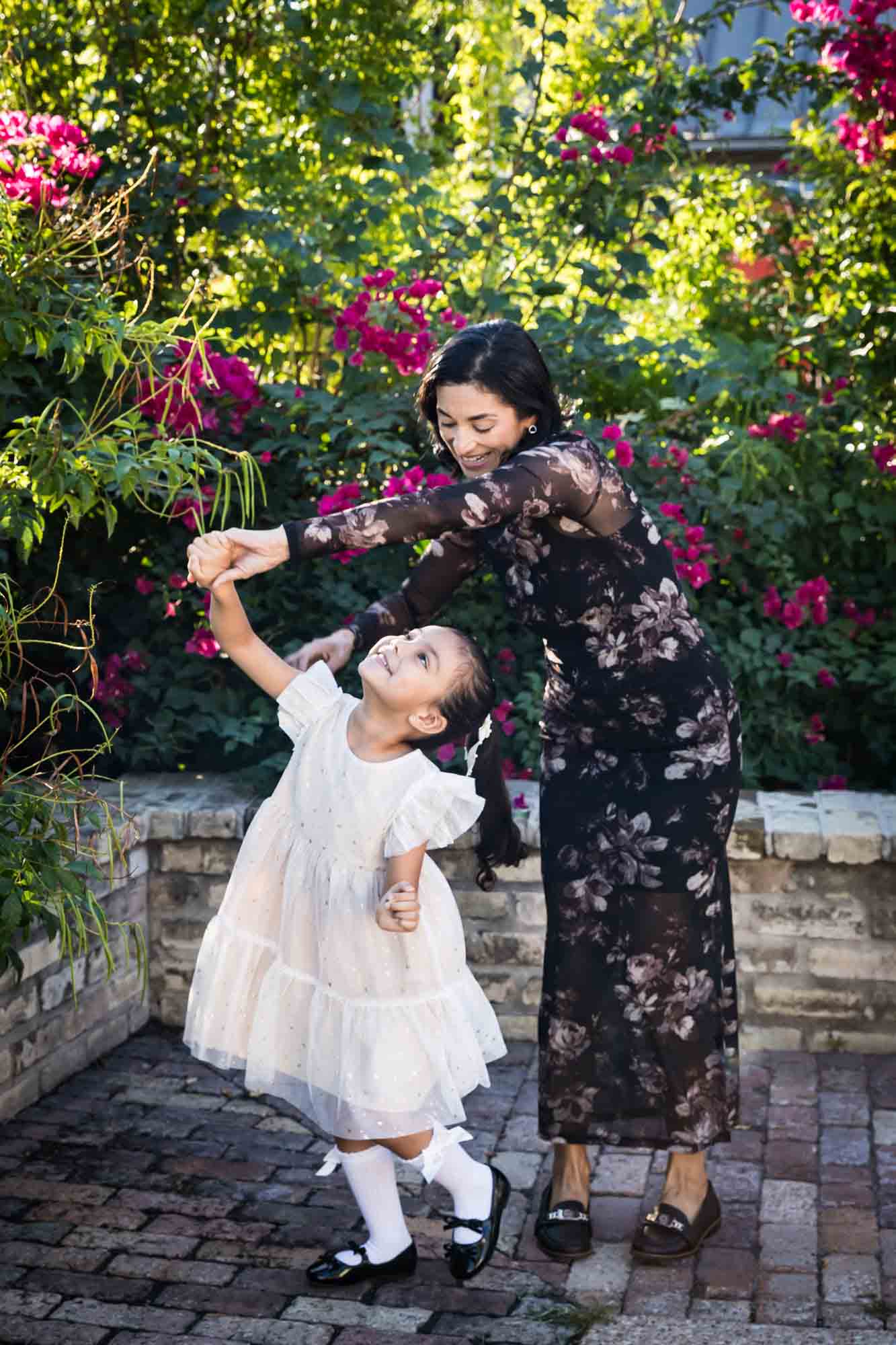 Mother dancing with little girl wearing white dress in front of brick wall and bushes during a La Villita family portrait session