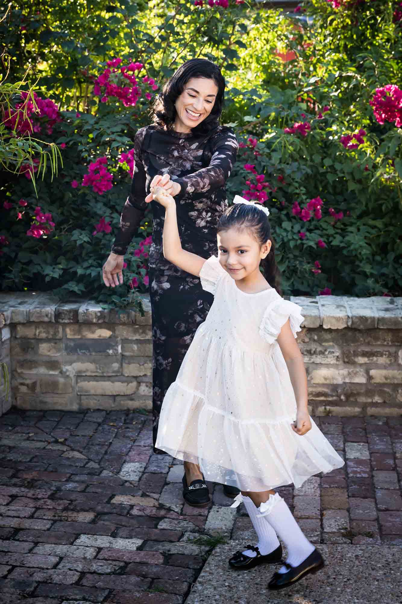 Mother dancing with little girl wearing white dress in front of brick wall and bushes during a La Villita family portrait session