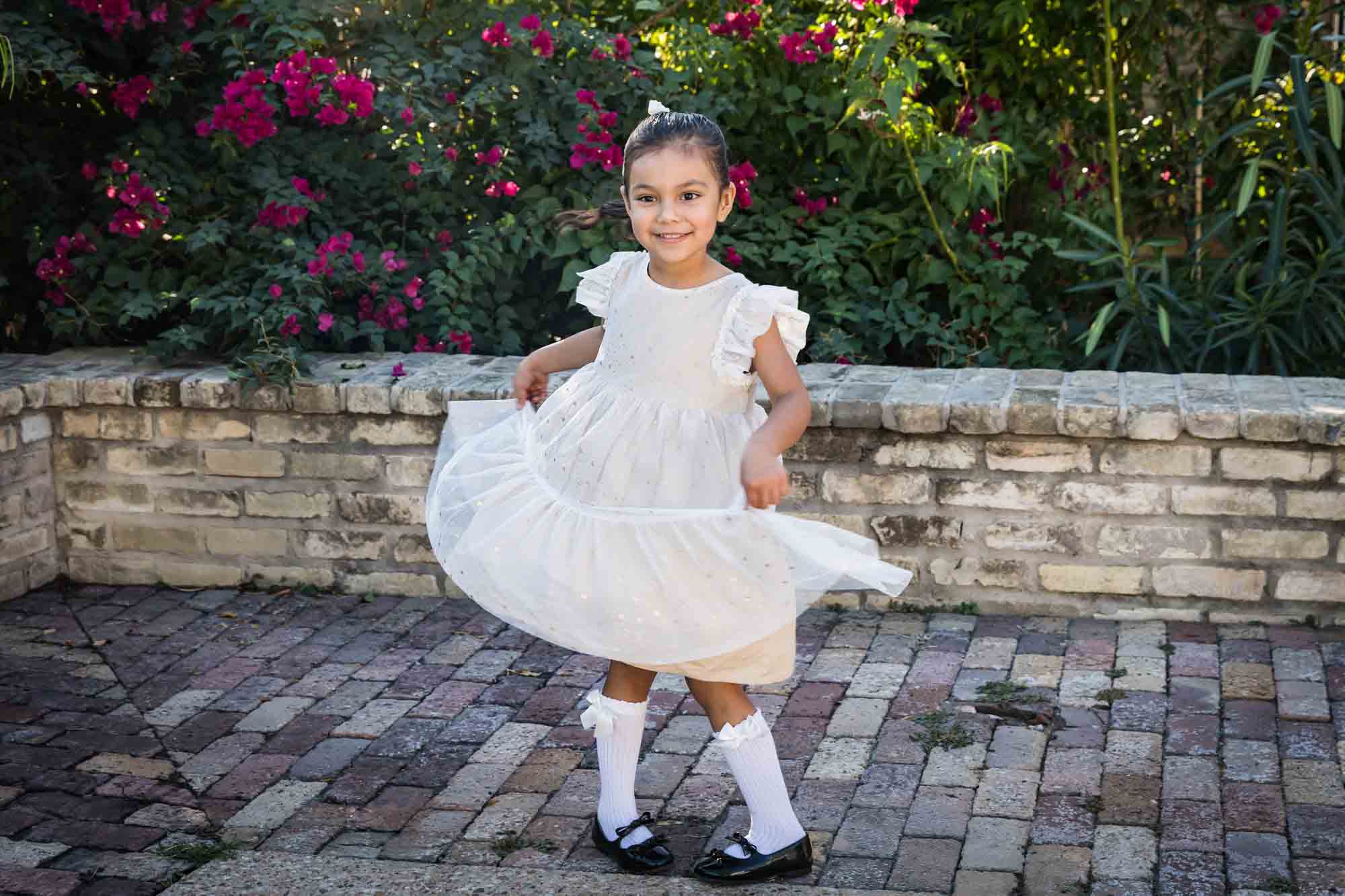 Little girl wearing white dress holding skirt in front of brick wall and bushes during a La Villita family portrait session