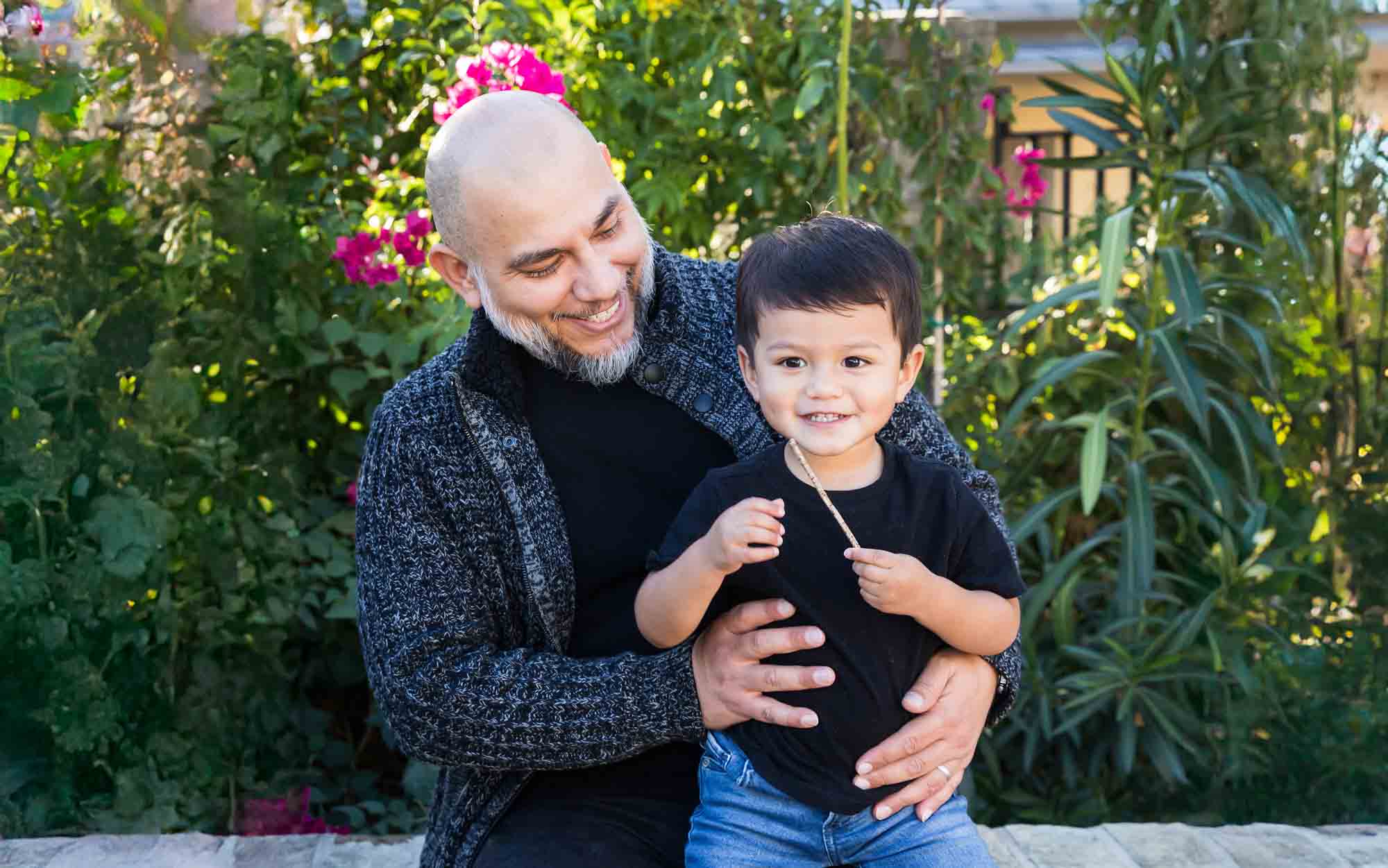 Father hugging little boy wearing jeans and black t-shirt in front of bushes during a La Villita family portrait session