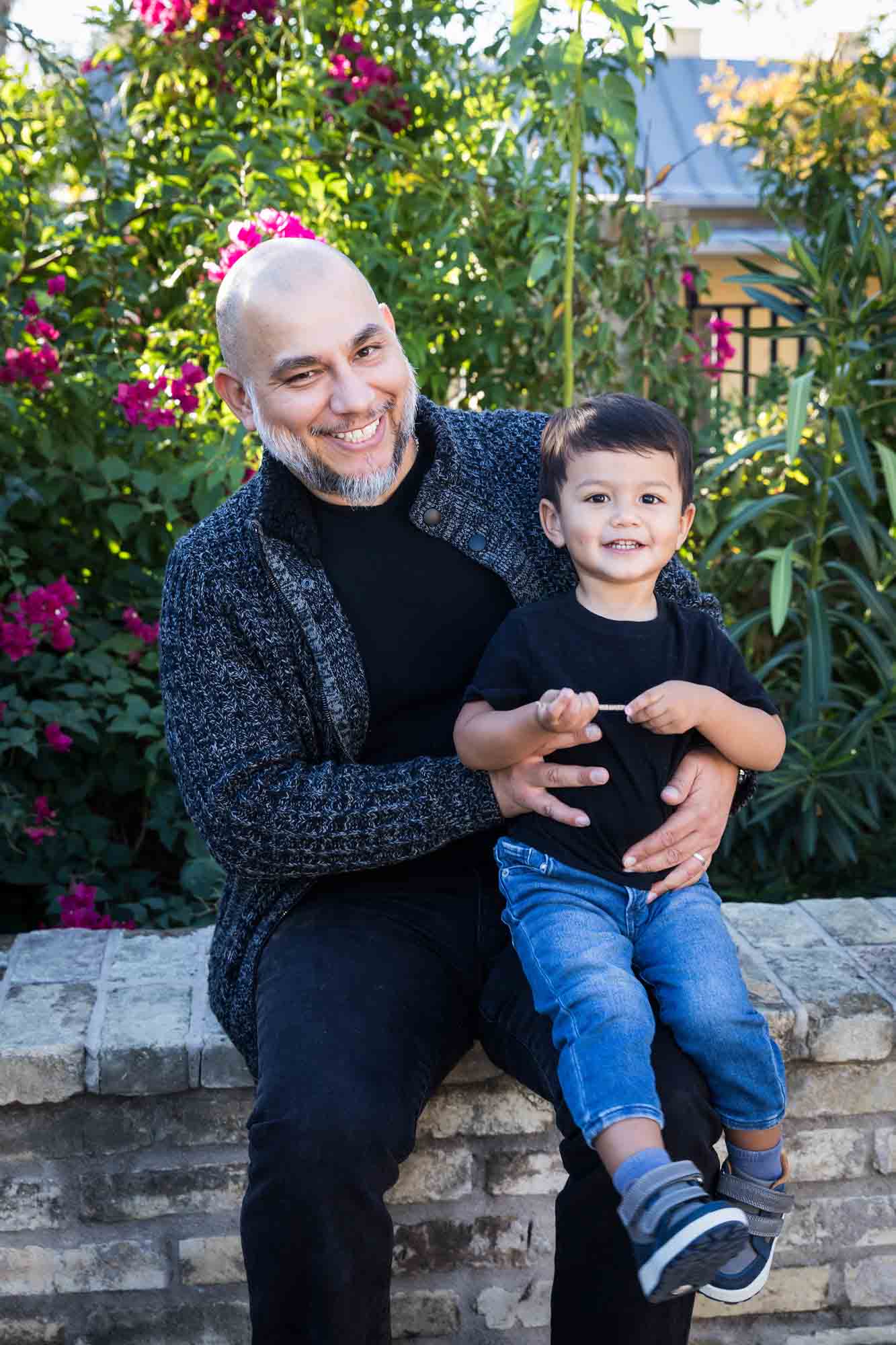 Father hugging little boy wearing jeans and black t-shirt in front of bushes during a La Villita family portrait session