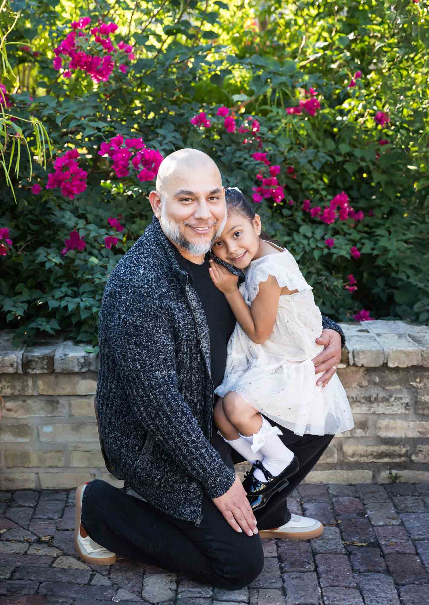 Father hugging little girl wearing white dress in front of bushes during a La Villita family portrait session