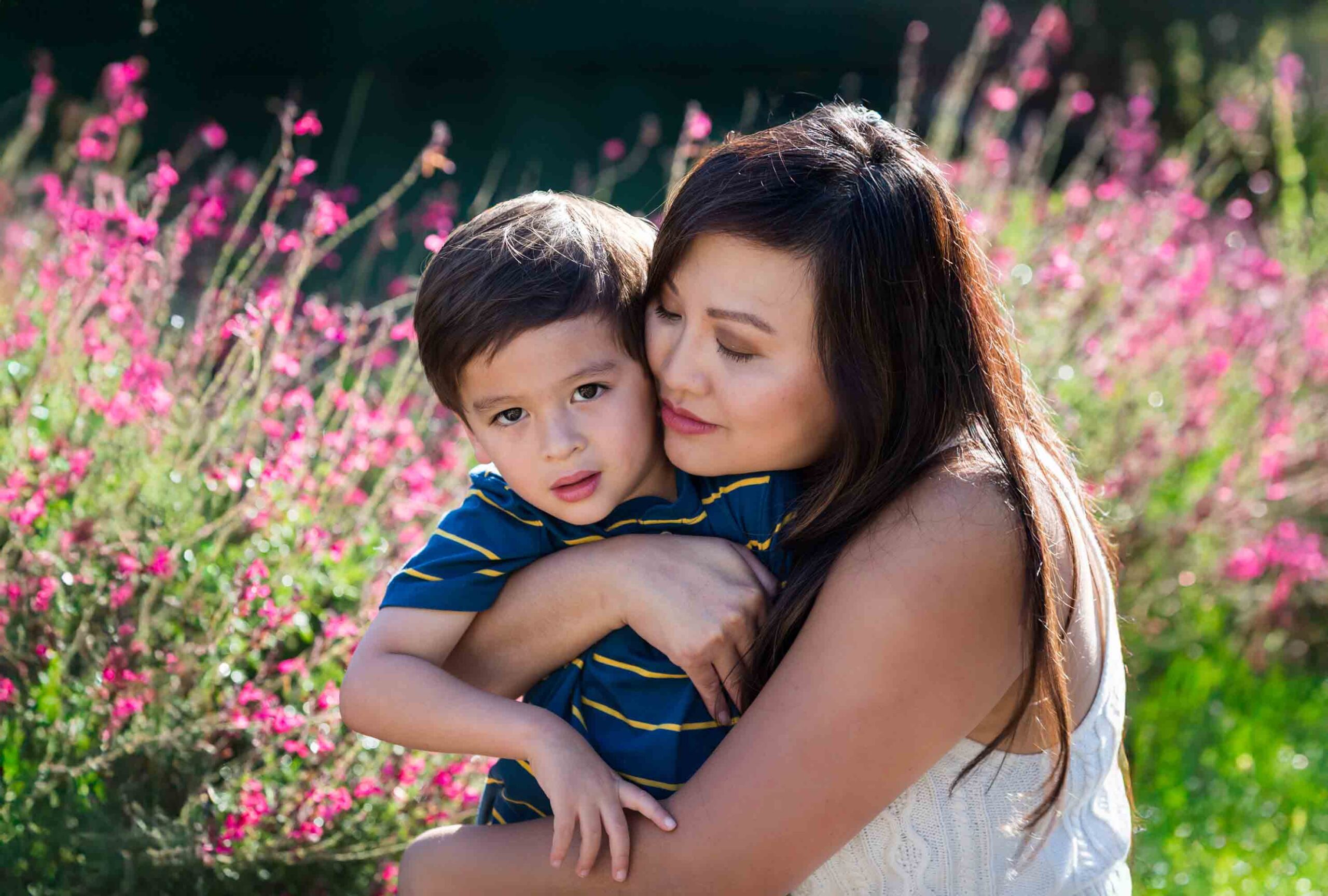 Mother hugging little boy wearing striped shirt in front of pink flowers for an article on photo tips for families with young children