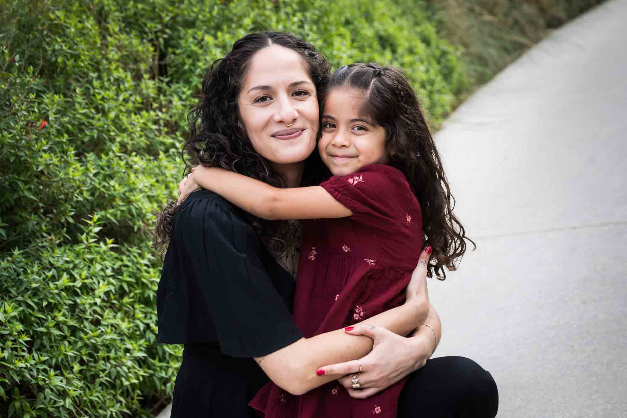 Mother and daughter hugging on sidewalk in front of green bushes during a San Pedro Creek Park family portrait session