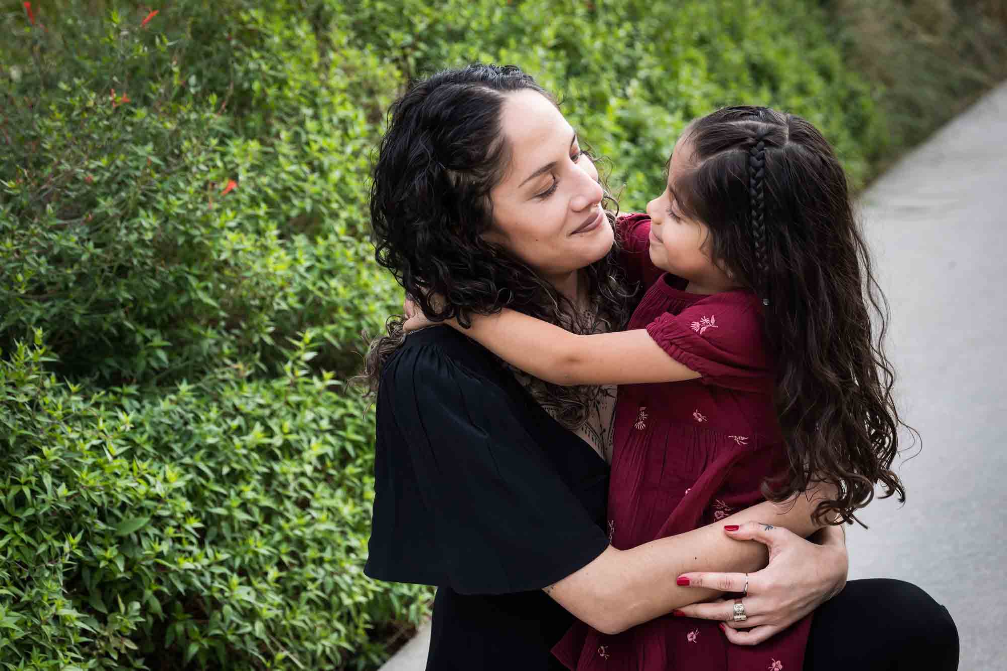 Mother and daughter hugging on sidewalk in front of green bushes during a San Pedro Creek Park family portrait session