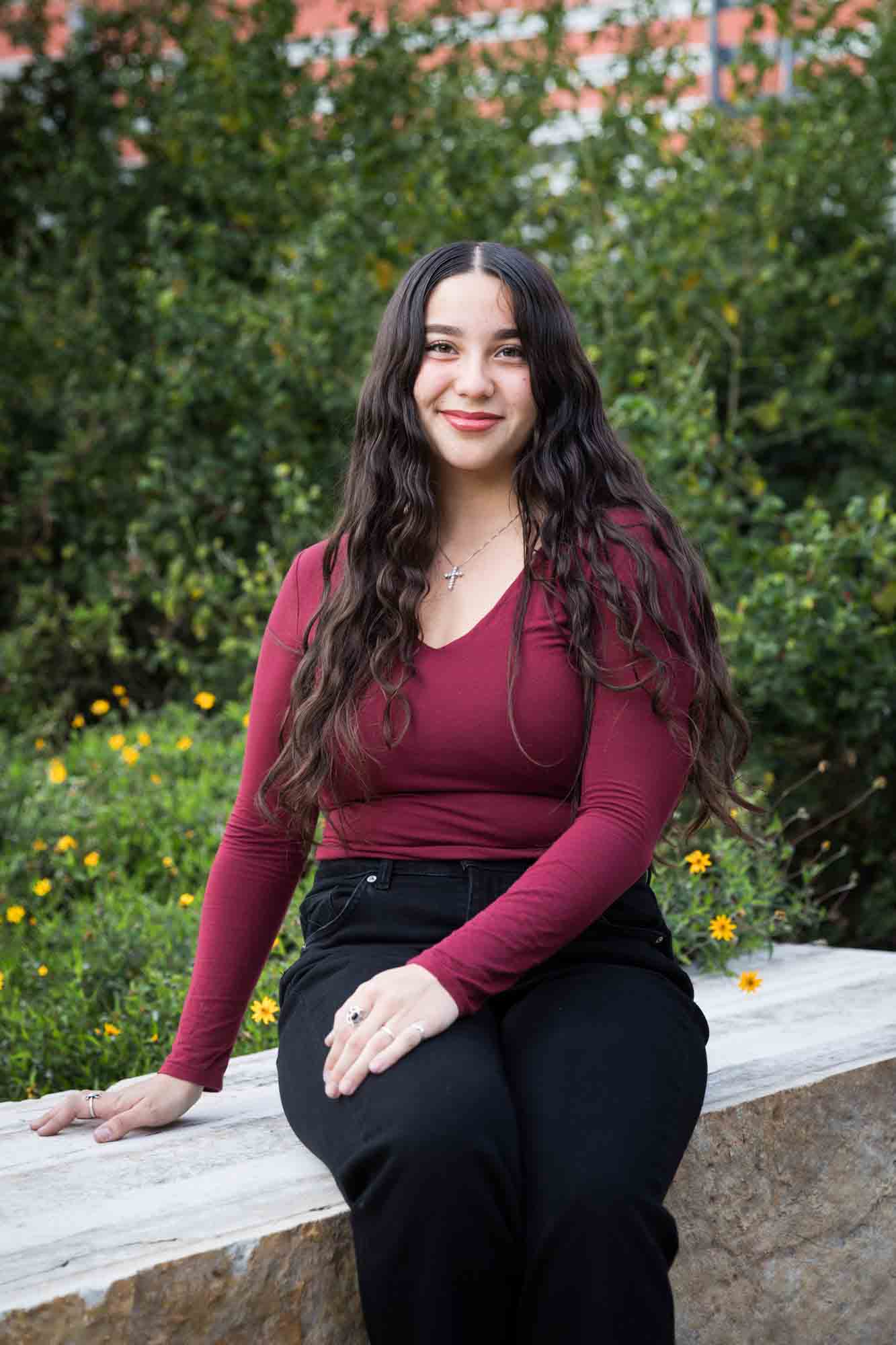 Teen girl with long brown hair wearing maroon top sitting in front of green bushes during a San Pedro Creek Park family portrait session