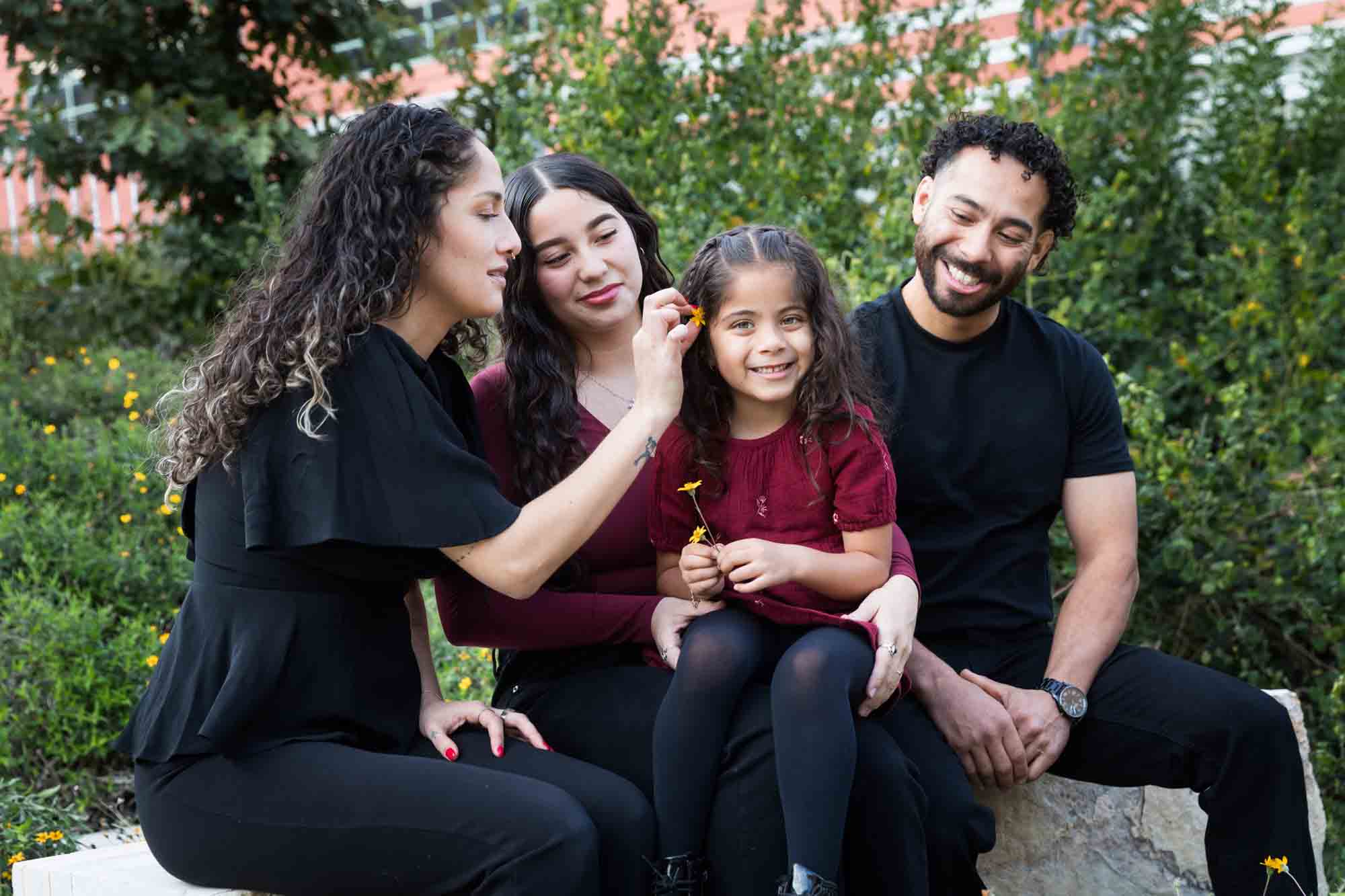 Parents, teen girl and little girl sitting on stone bench playing with flower during a San Pedro Creek Park family portrait session