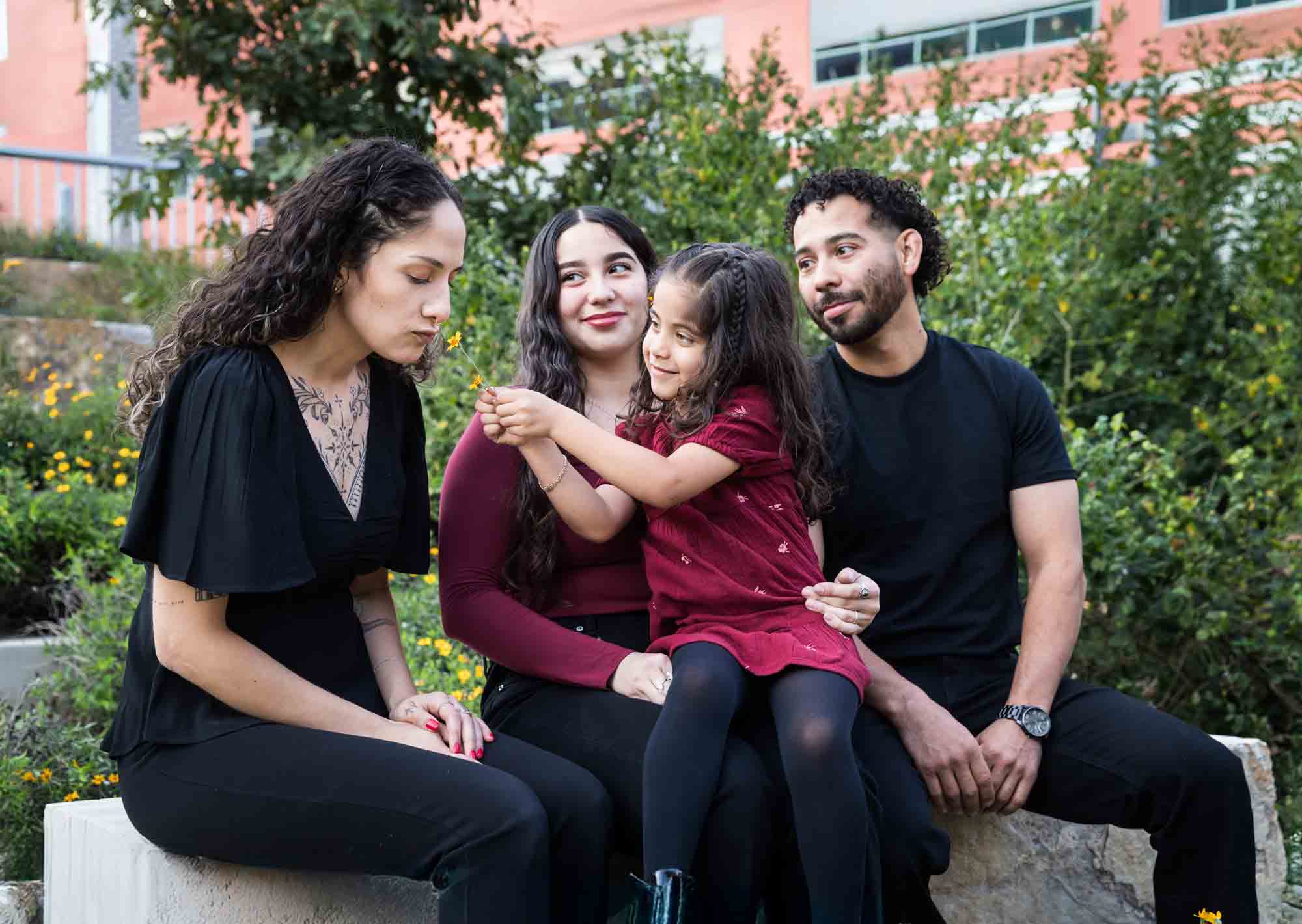 Parents, teen girl and little girl sitting on stone bench playing with flower during a San Pedro Creek Park family portrait session