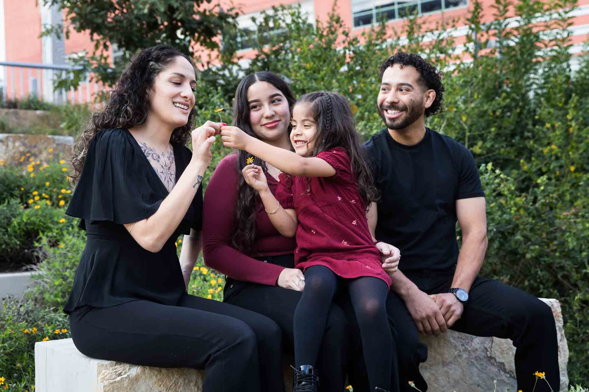 Parents, teen girl and little girl sitting on stone bench playing with flower during a San Pedro Creek Park family portrait session