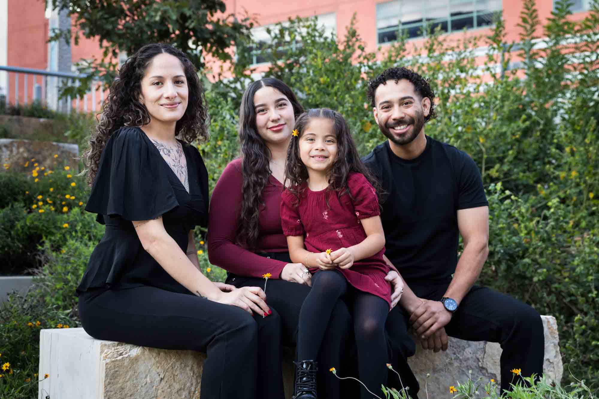 Parents, teen girl and little girl sitting on stone bench playing with flower during a San Pedro Creek Park family portrait session