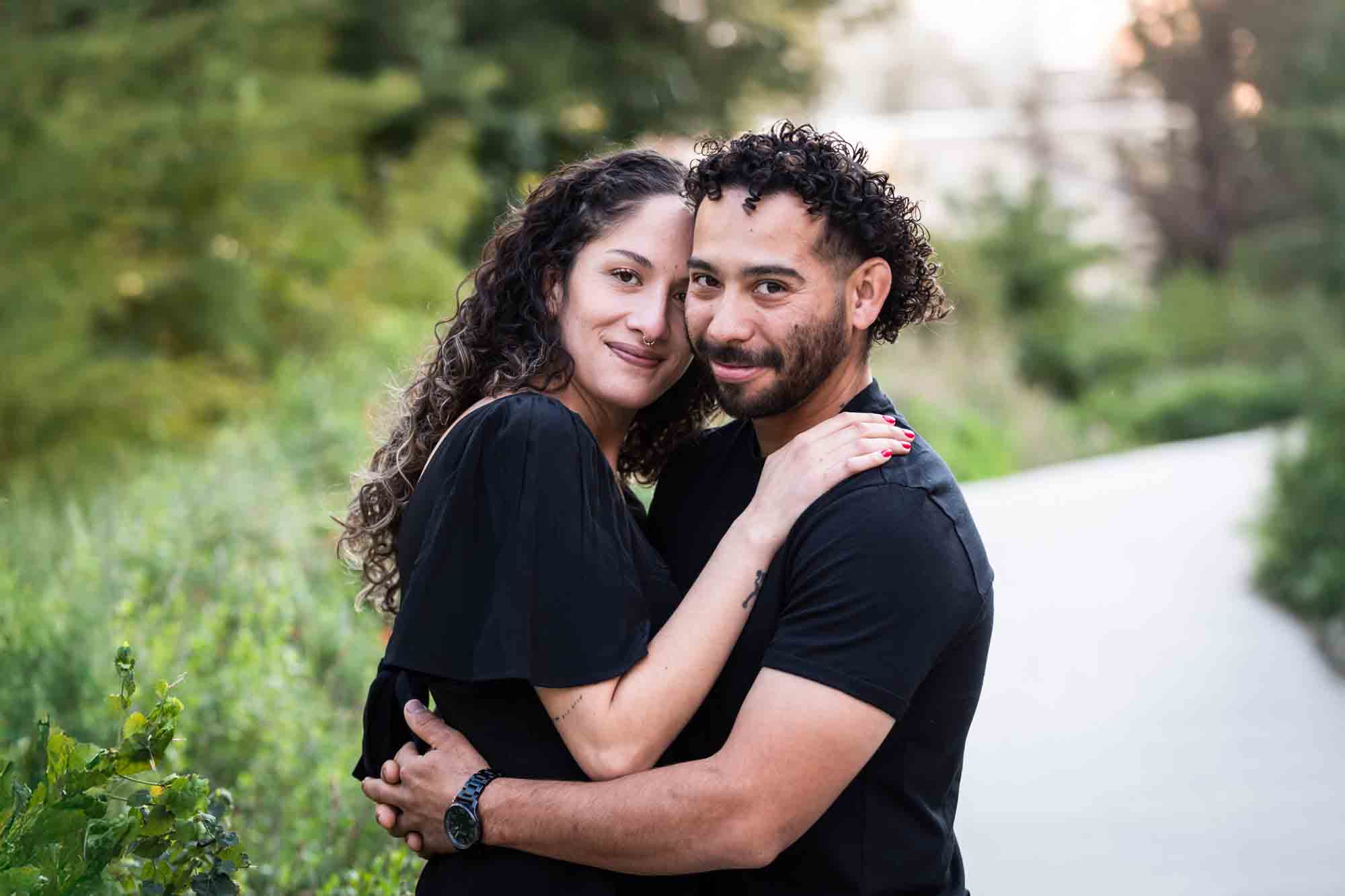 Couple wearing black t-shirts hugging in front of green bushes during a San Pedro Creek Park family portrait session