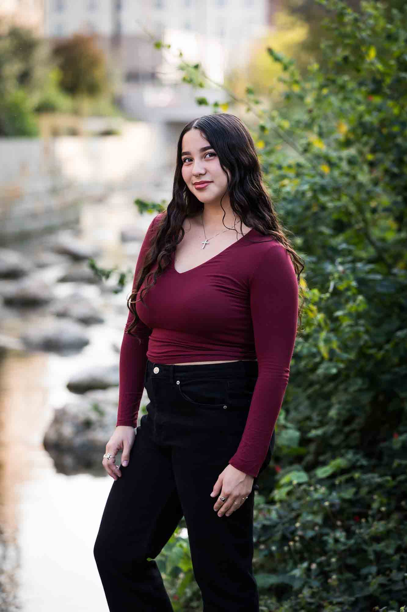 Teen girl with long brown hair wearing maroon top standing in front of green bushes and river during a San Pedro Creek Park family portrait session