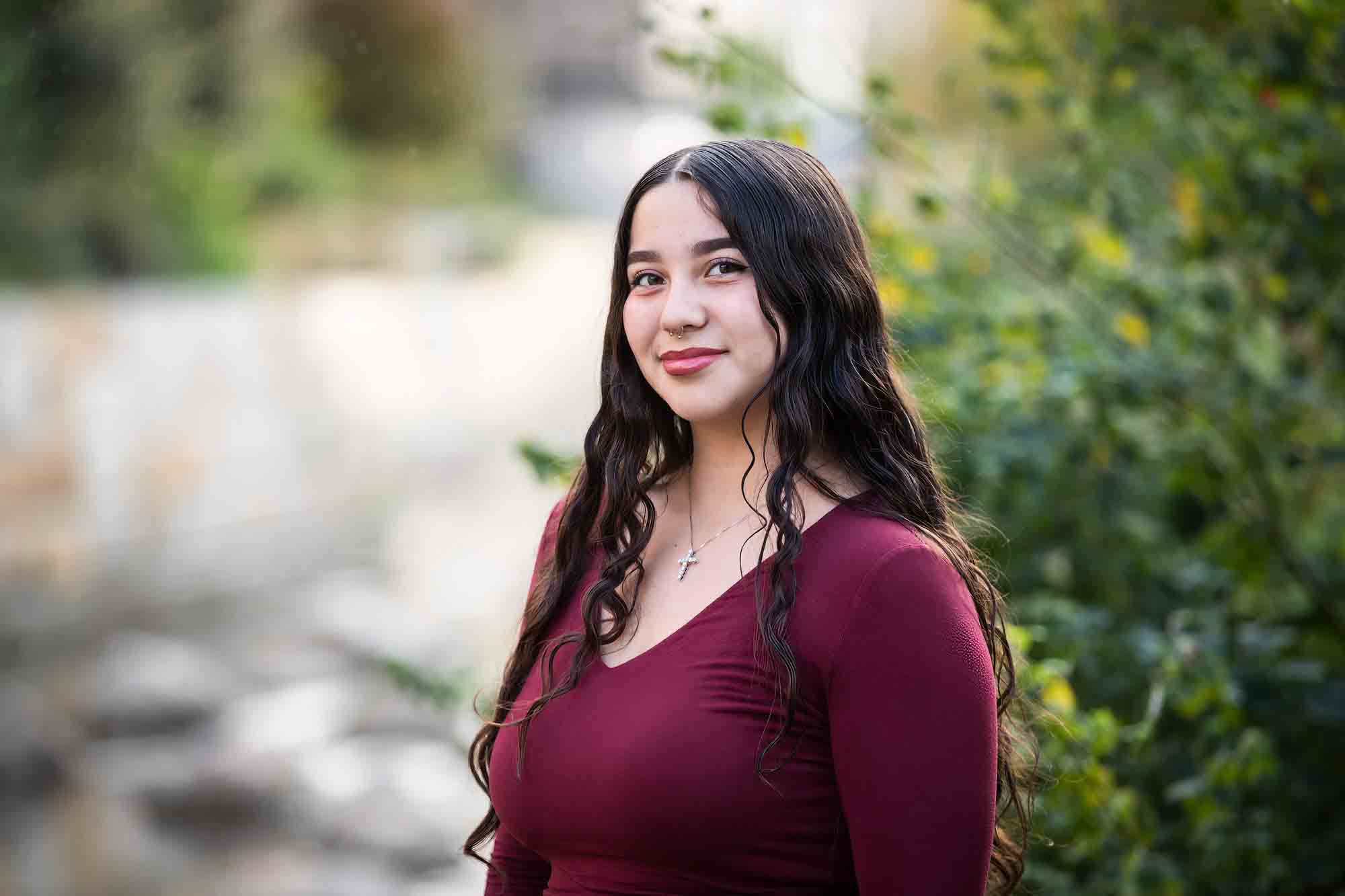 Teen girl with long brown hair wearing maroon top standing in front of green bushes during a San Pedro Creek Park family portrait session