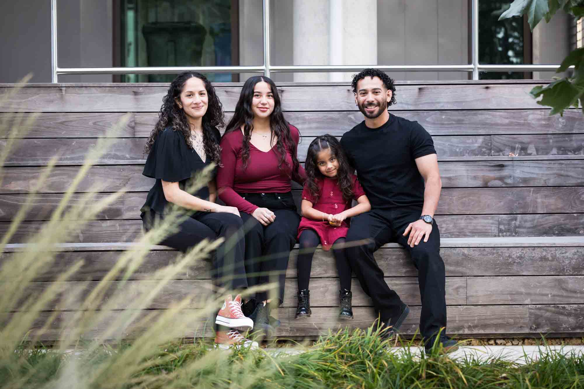 Family of two parents, teen girl and little girl sitting on wooden benches by UTSA Data Sciences building