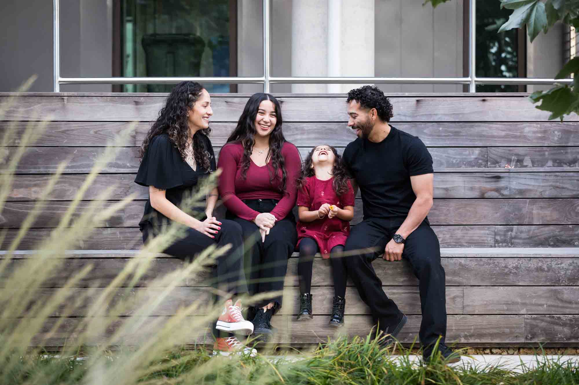 Family of two parents, teen girl and little girl sitting on wooden benches by UTSA Data Sciences building