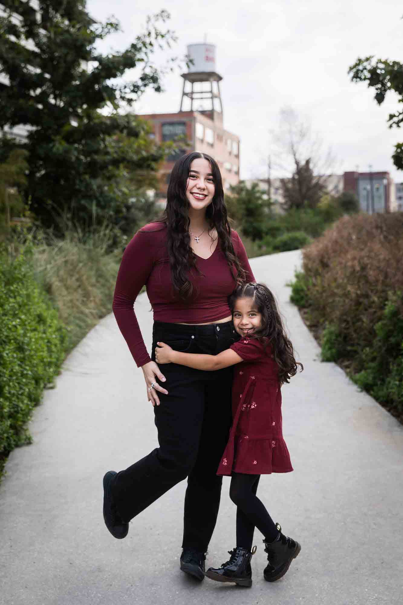 Older and younger sisters wearing maroon clothes hugging on sidewalk during a San Pedro Creek Park family portrait session