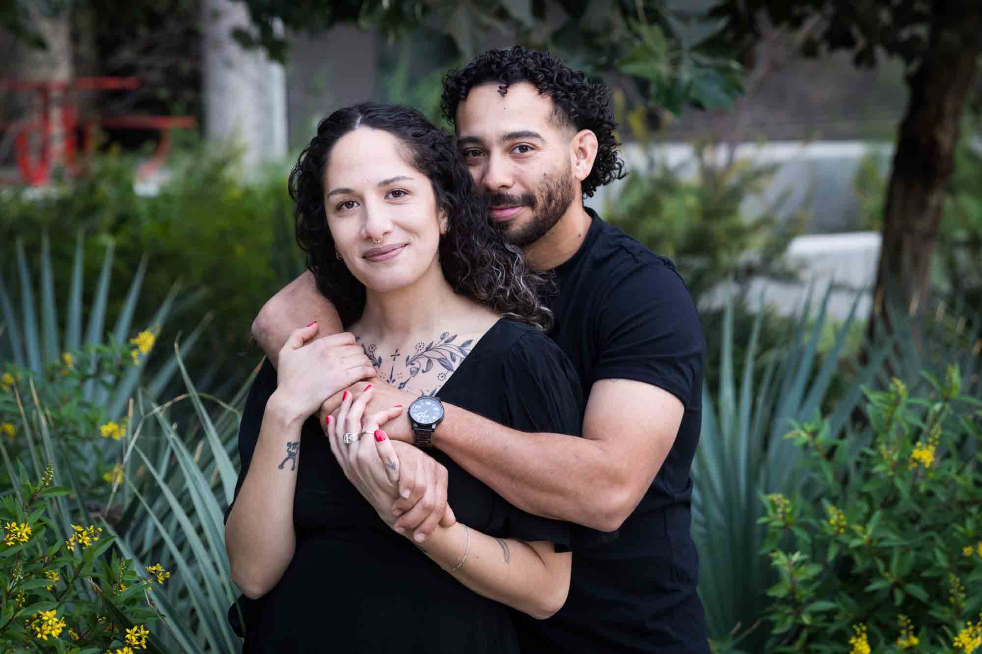 Couple wearing black t-shirts hugging in front of green bushes and palm fronds