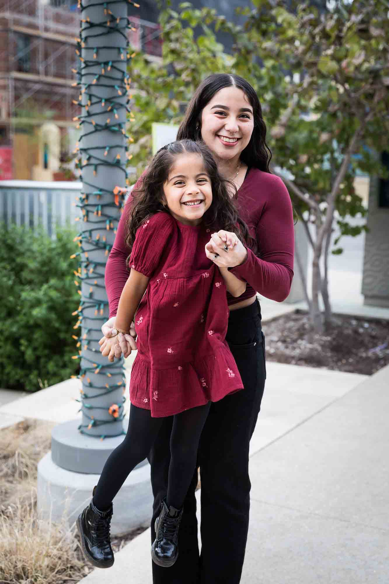 Older and younger sisters wearing maroon clothes holding hands on sidewalk