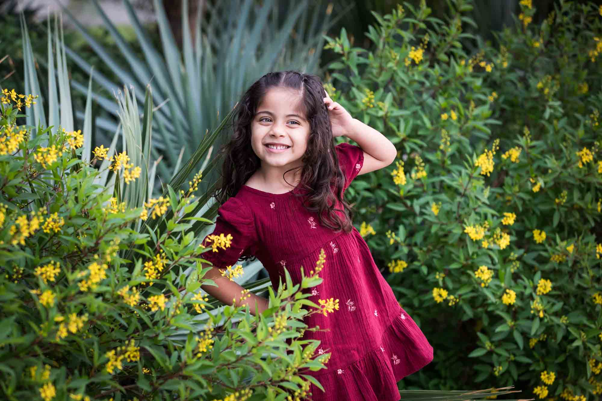 Little girl with long brown hair wearing maroon dress standing in front of green bushes